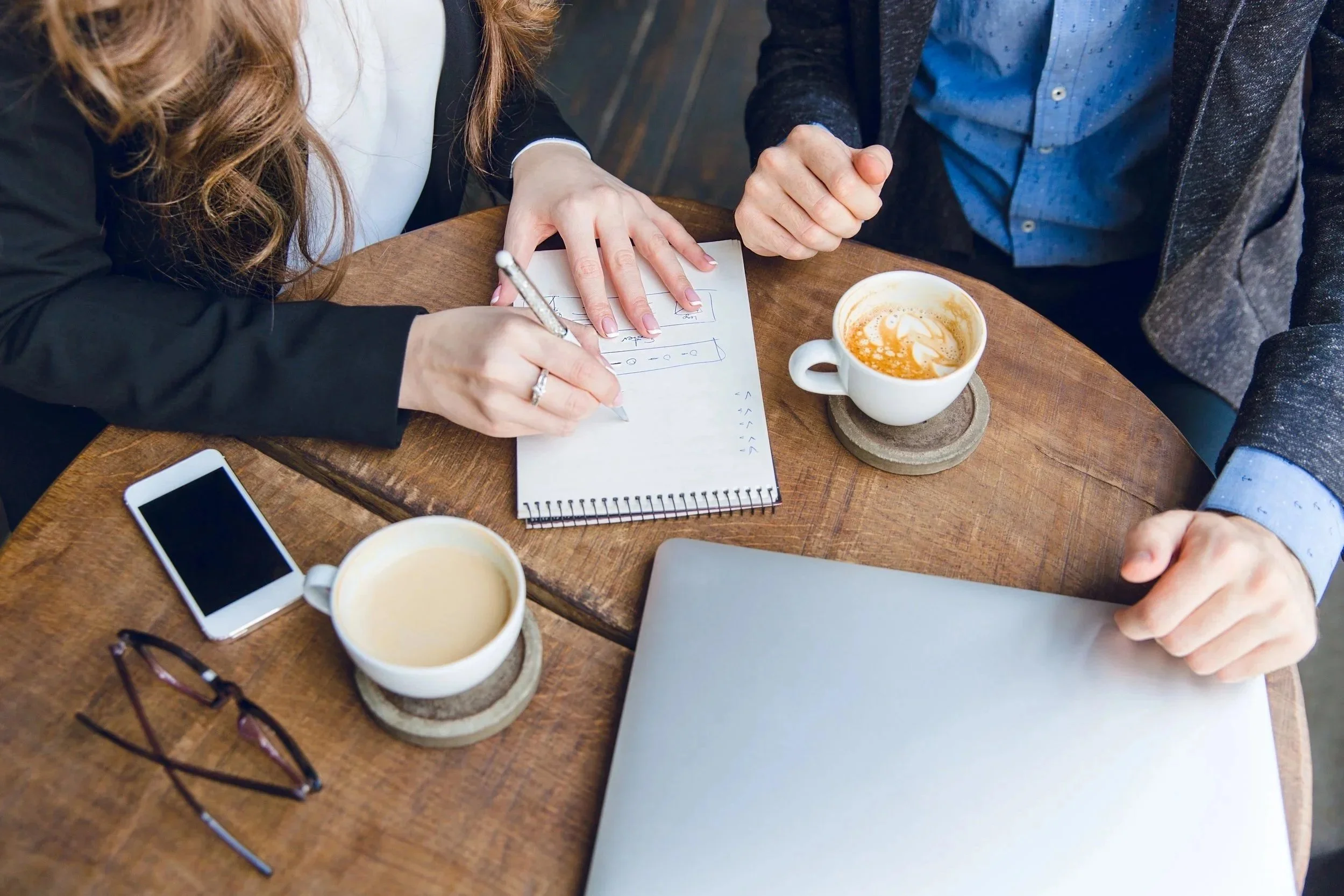 Two people sitting at a wooden table, one writing on a notepad with handwritten notes and diagrams, the other pointing at the notepad. There are two cups of coffee, a smartphone, a pair of glasses, and a laptop on the table.