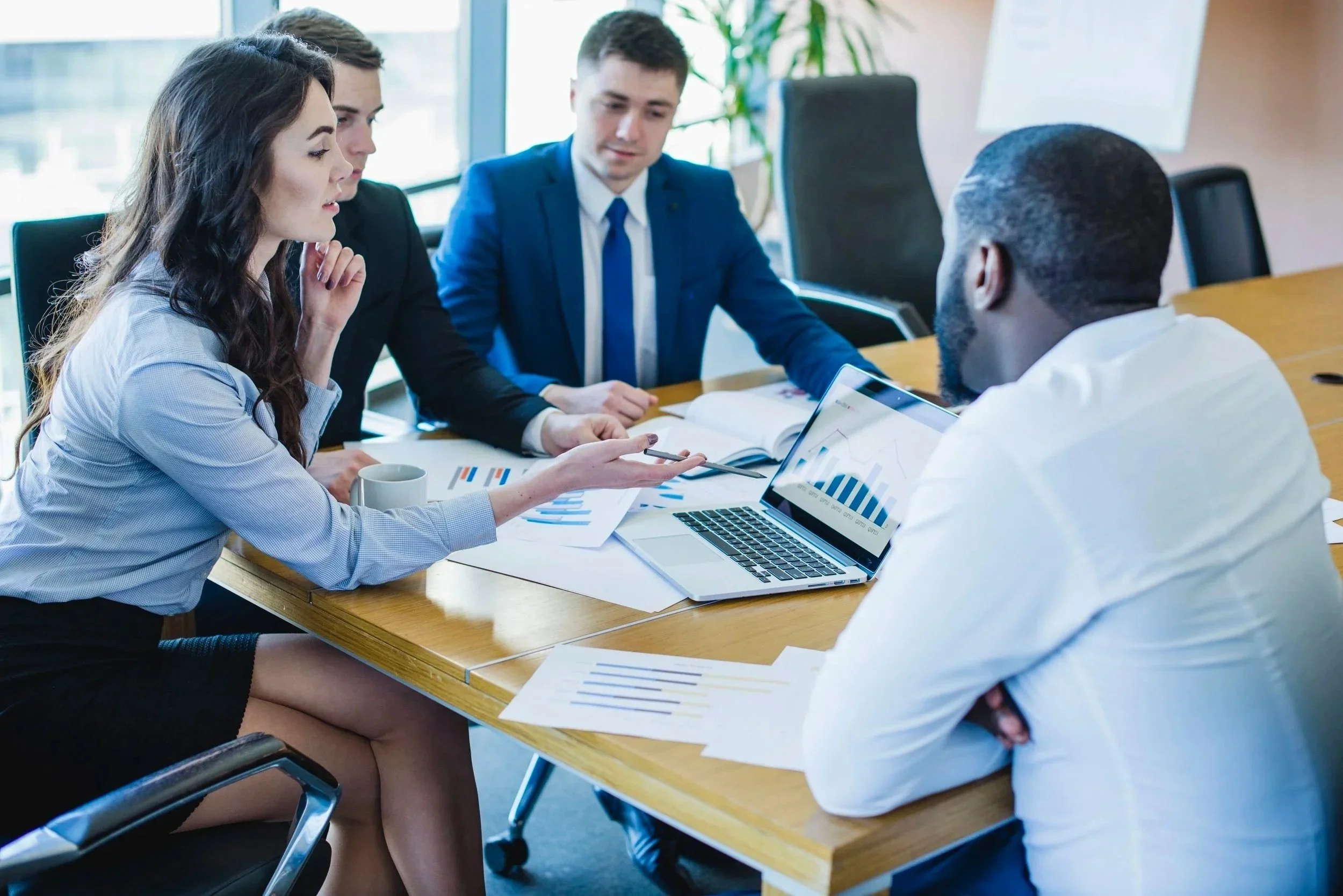 Five business professionals, three women and two men, are sitting around a wooden conference table engaged in a discussion. They are looking at printed documents, a laptop displaying charts, and a tablet. The setting is a modern office with large windows and a plant in the background.