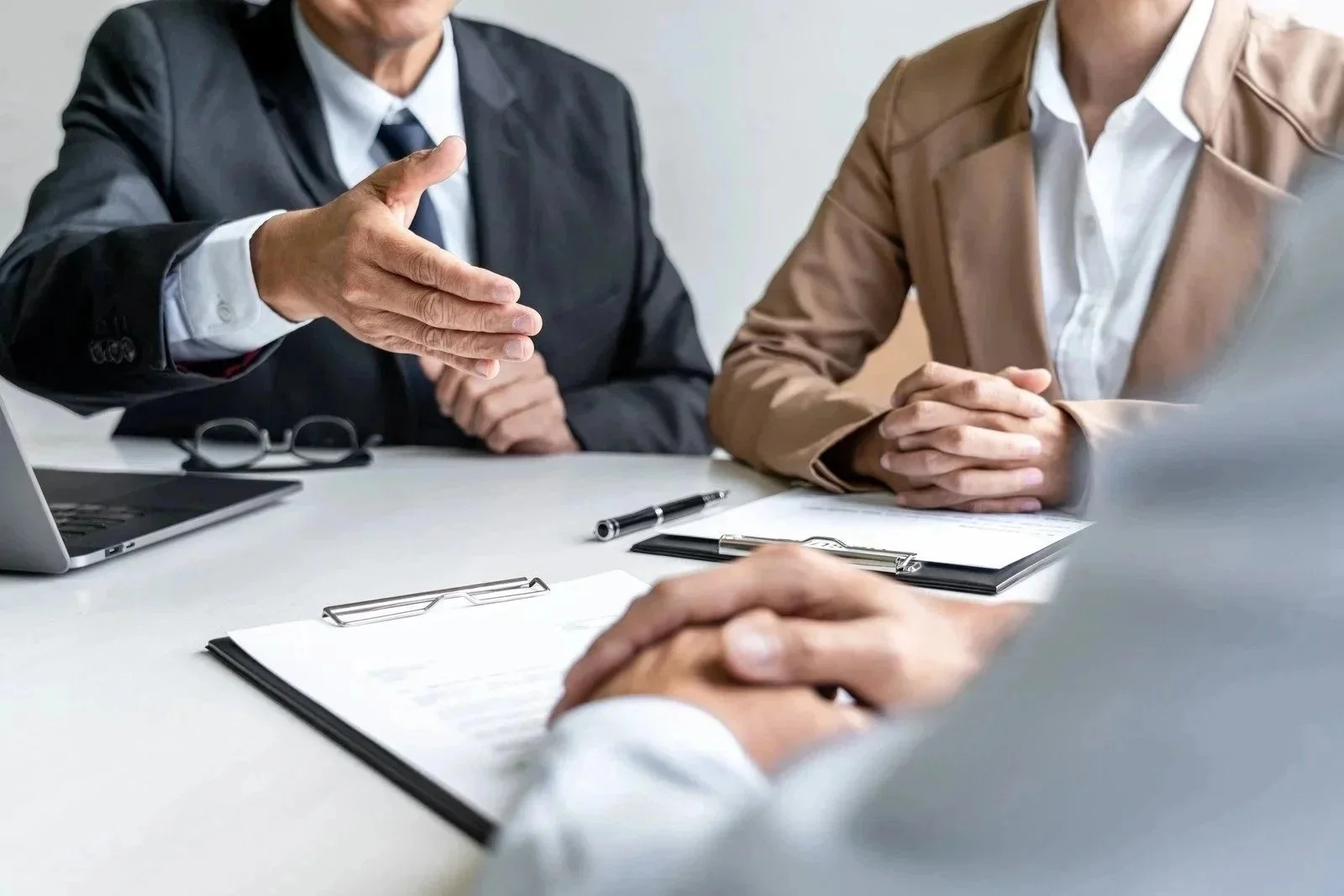 Business meeting with three people in suits, one person gesturing with an open hand, documents, pens, and a laptop on the table.