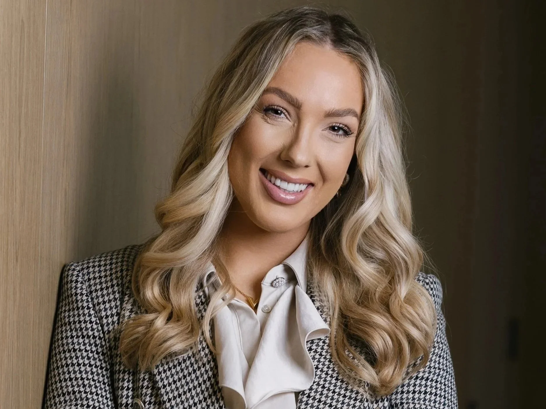 A young woman with long blonde wavy hair, smiling, dressed in a dark blazer and white blouse with ruffles, standing indoors next to a wooden wall and green foliage.
