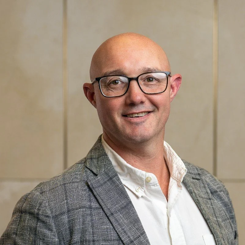 A man with a bald head and glasses smiling, wearing a white shirt and a grey checked blazer, standing against a beige background.