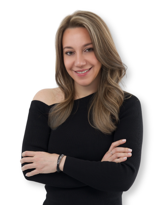 A young woman with long, wavy blonde hair, smiling and looking at the camera, wearing a black off-the-shoulder long-sleeve top, with her arms crossed.