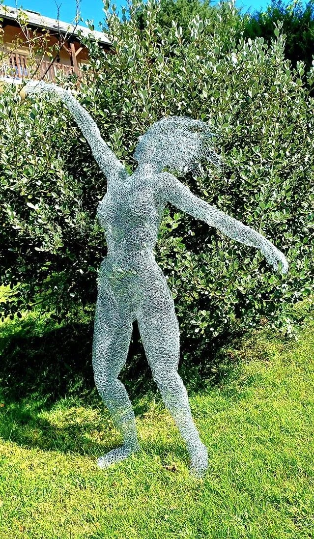 Une sculpture métallique représentant une femme nue avec les bras levés, placée devant un buisson vert dans un jardin.