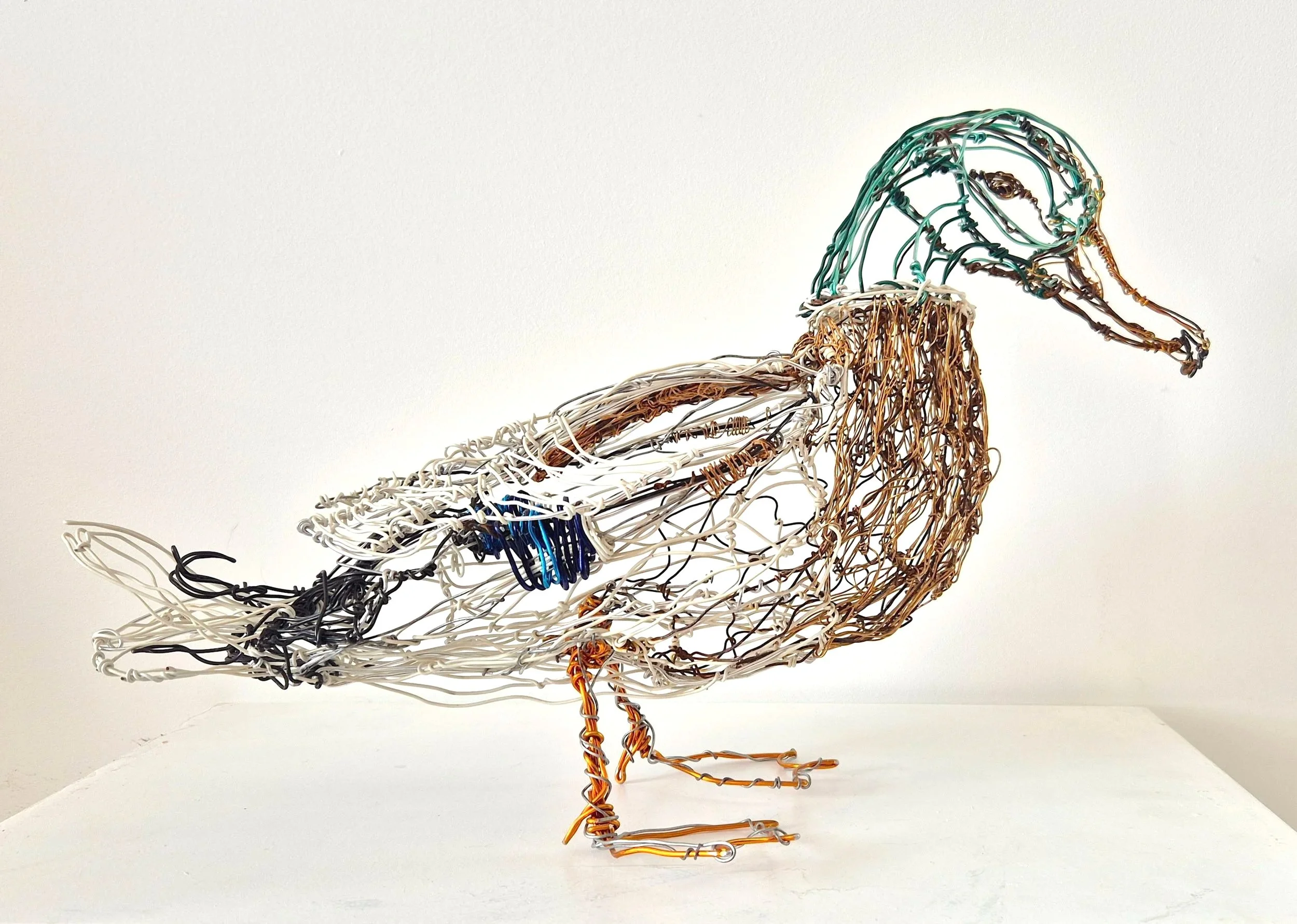 Une sculpture en fil de métal coloré représentant un flamant rose. La sculpture est placée sur un fond blanc, avec un socle simple, mettant en valeur les détails et les couleurs de l'oiseau.