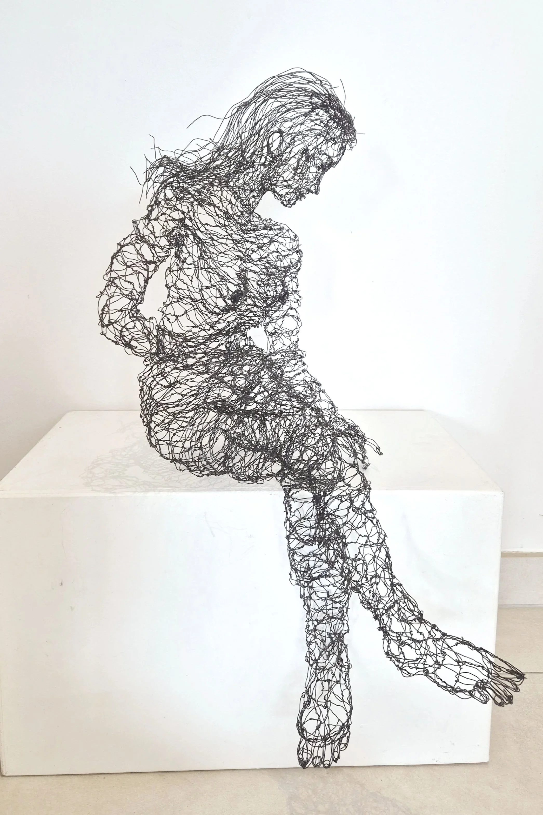 Sculpture en fil métallique représentant une femme assise, regardant vers le bas, sur un socle blanc, fond blanc