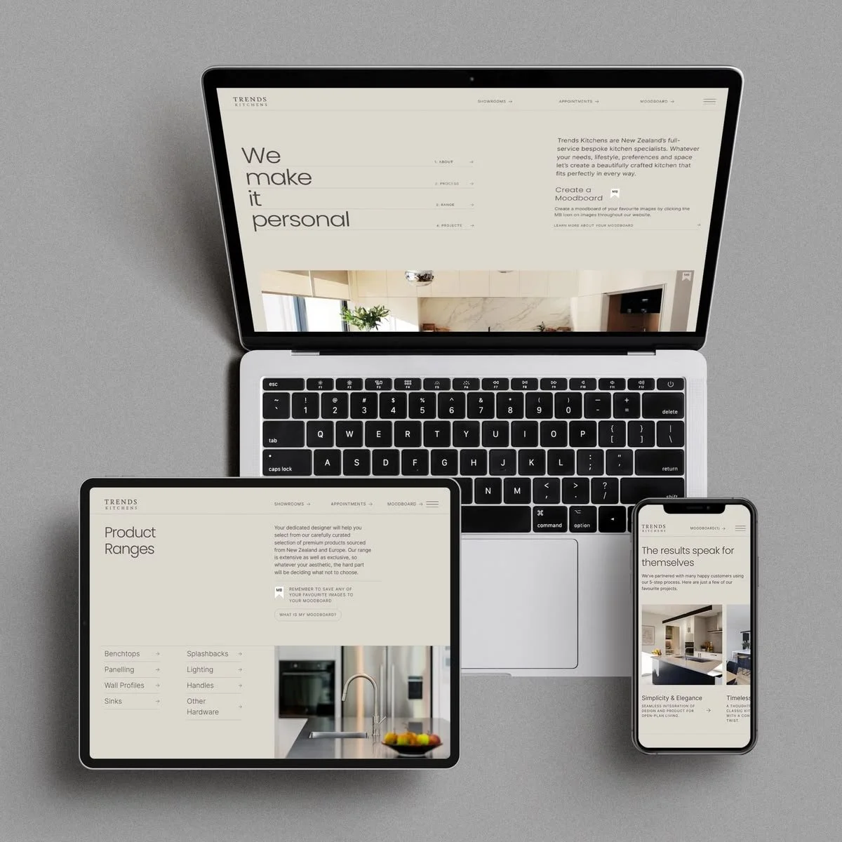 A laptop, tablet, and smartphone showing a website for Trends Kitchens, a kitchen design company, on a gray surface.
