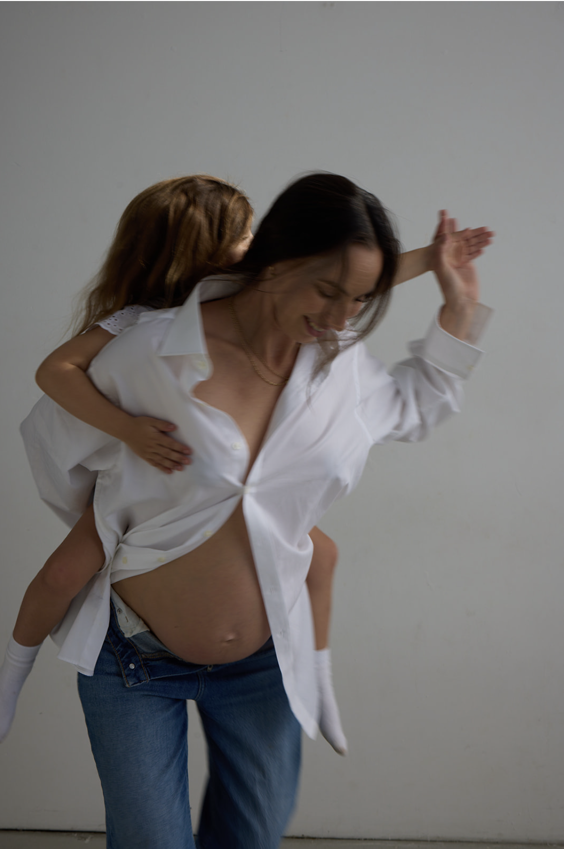 A woman with dark hair is carrying a young girl on her back. The woman is wearing a partially unbuttoned white shirt and jeans, and the girl is wearing a white top with eyelet sleeves. The woman is smiling and appears happy.