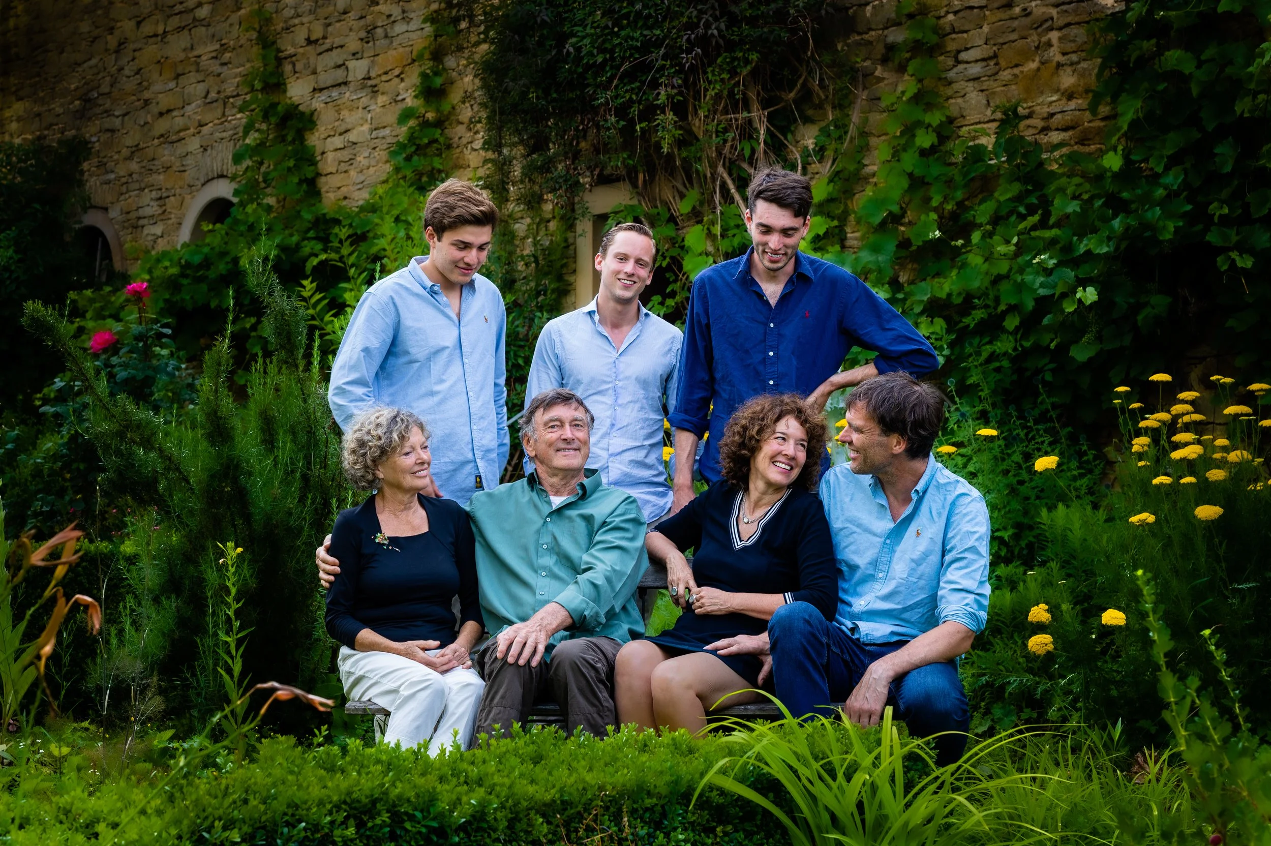 Eine Gruppe von acht Menschen unterschiedlichen Alters sitzt und steht in einem Garten voller Grünpflanzen und Blumen vor einer Steingartenwand. Sie scheinen sich locker zu unterhalten und zu amüsieren.