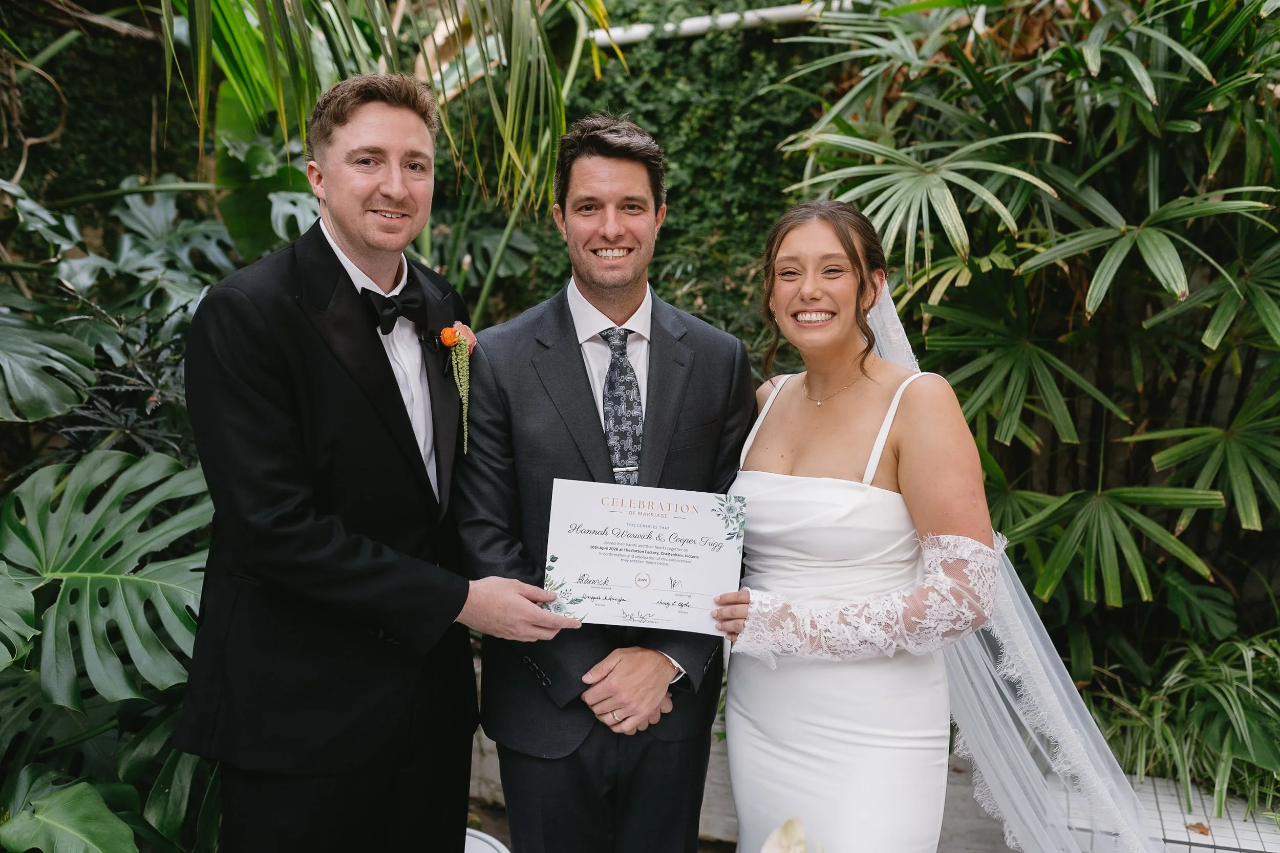 A bride and groom at their wedding ceremony, holding a certificate with a man in a suit standing between them, all smiling, surrounded by lush green tropical plants.