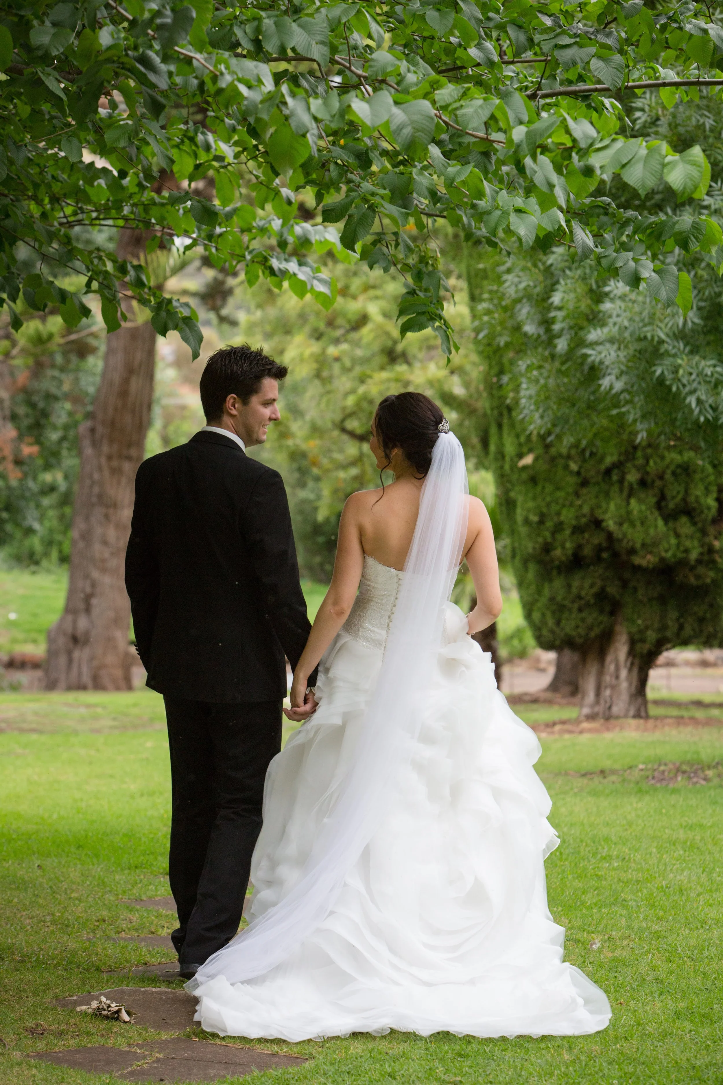 A bride and groom holding hands outdoors, walking on a stone pathway in a park surrounded by green trees and grass.
