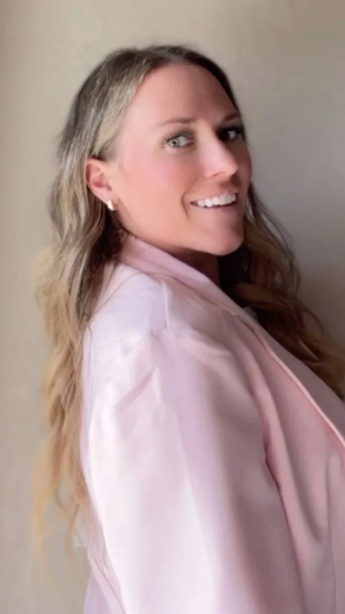 A woman with long, wavy blonde hair smiling and looking at the camera, wearing a light pink blazer, standing against a plain beige wall.