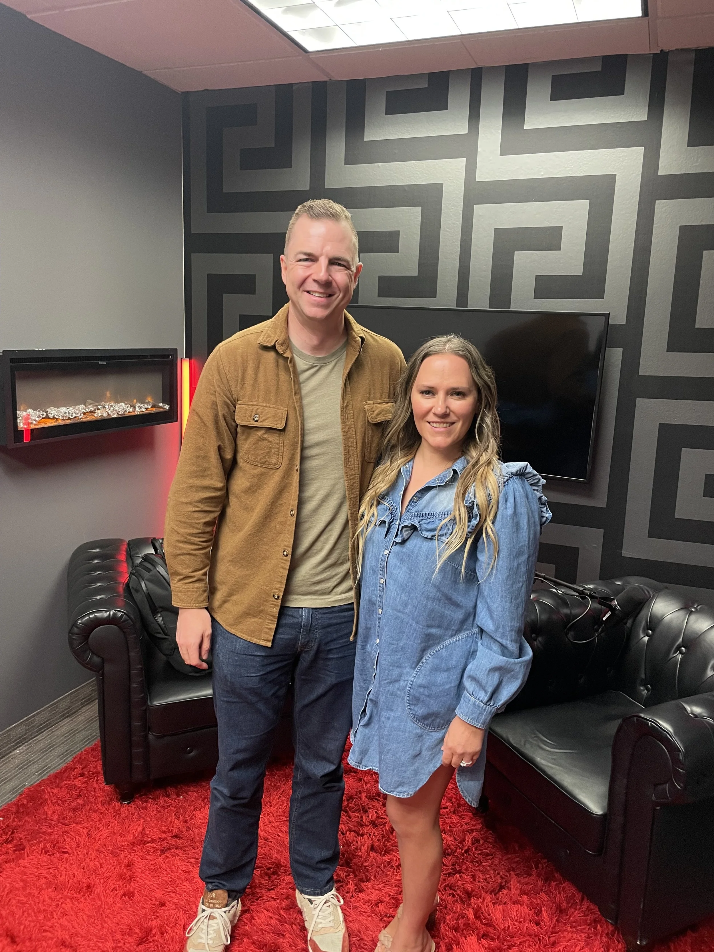 A man and woman are standing together in a modern room with a black patterned wall, a television, a black leather couch, and a red shag rug. The man is wearing a brown jacket and jeans, and the woman is wearing a denim dress. They are smiling and posing for the photo.