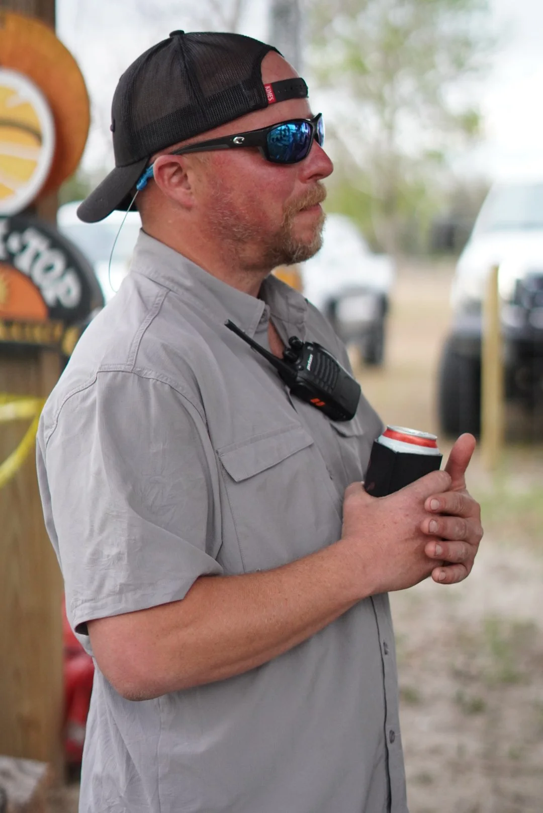 A man wearing sunglasses and a black trucker hat, holding a can of soda in a black koozie, standing outdoors with vehicles and trees in the background.