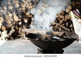 Charcoal grill with smoke rising, placed on the ground near stacked firewood.