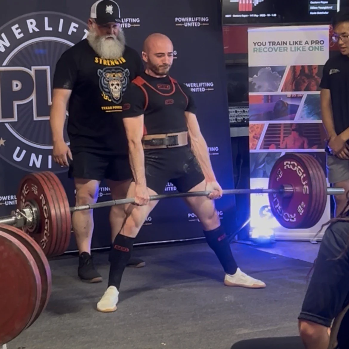 A male powerlifter in a black and red uniform is performing a deadlift during a competition, with an older man with a white beard and black cap supervising behind him. There is a woman standing to the right, observing. The setting is indoors, with banners in the background promoting powerlifting and fitness.