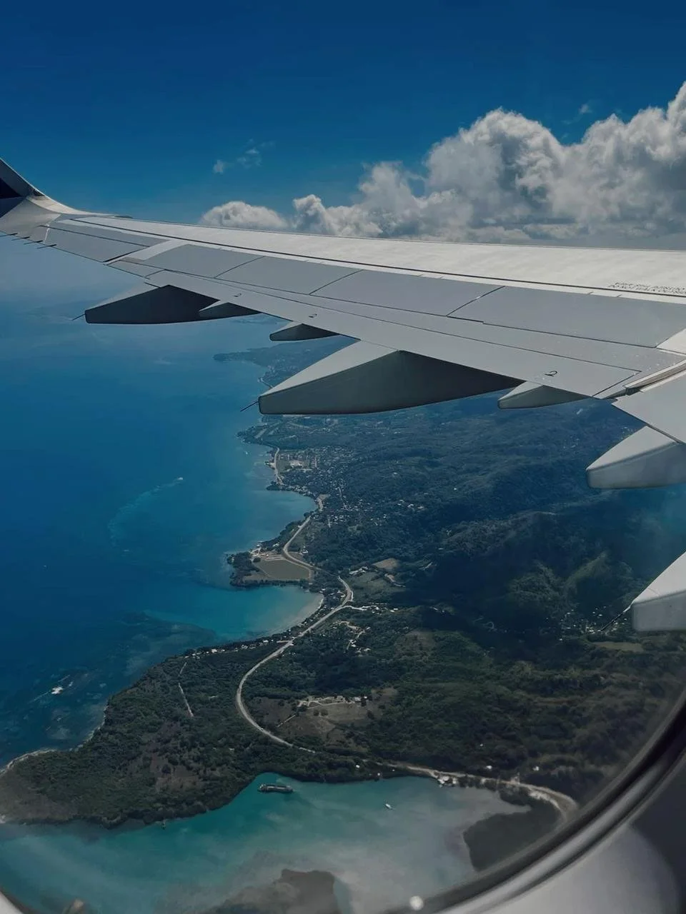 View from an airplane window showing coastline, ocean, and land with roads and greenery under a partly cloudy sky.