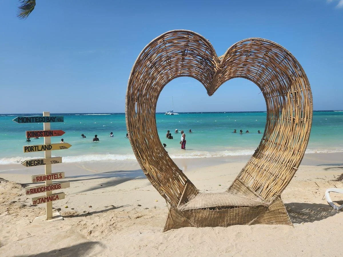 A beach scene with a wicker heart-shaped frame and a directional signpost pointing to various locations, with the ocean and people swimming in the background.