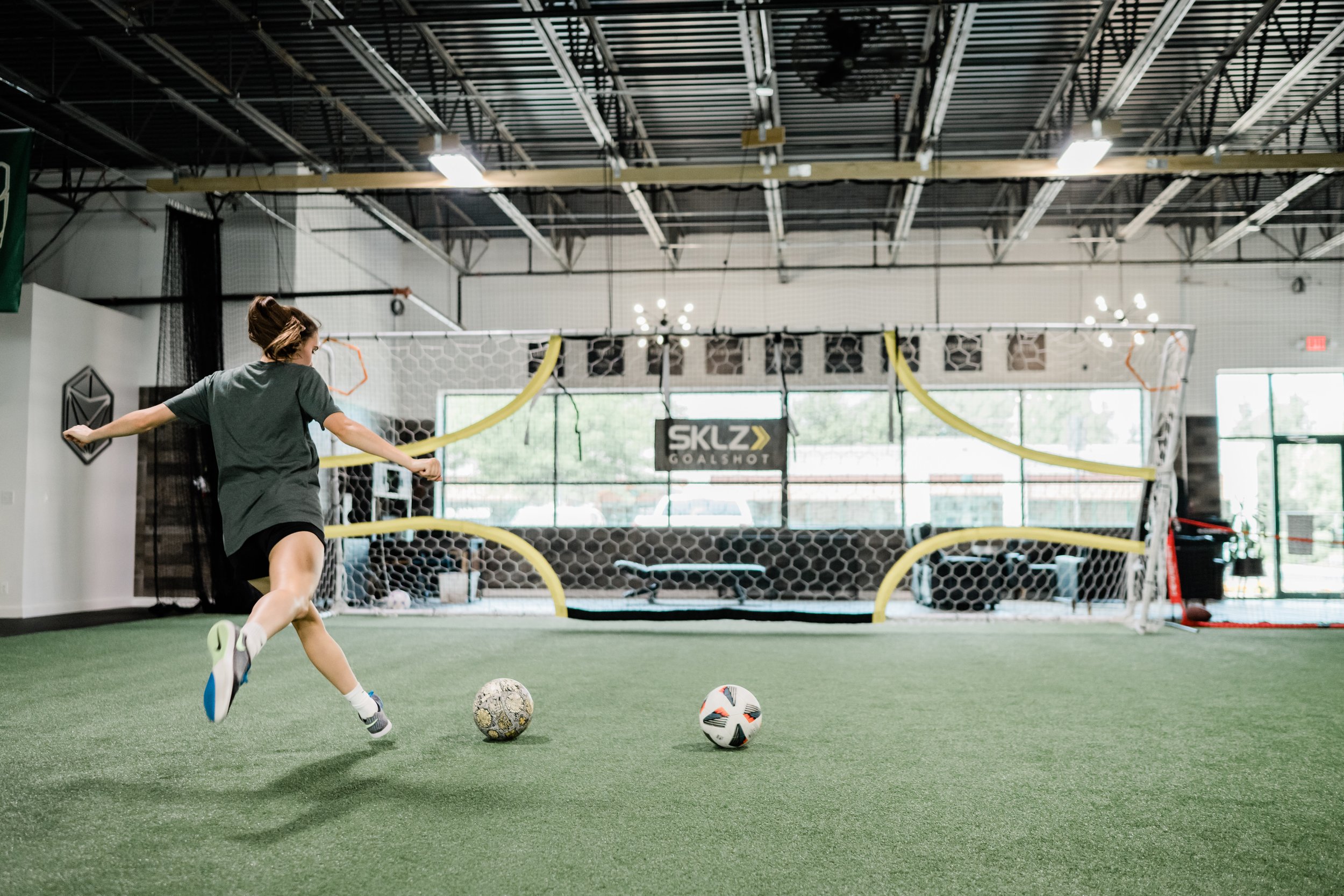 A female athlete practicing soccer indoors, kicking a soccer ball on artificial turf with multiple soccer balls in front of her, and a goal net in the background.