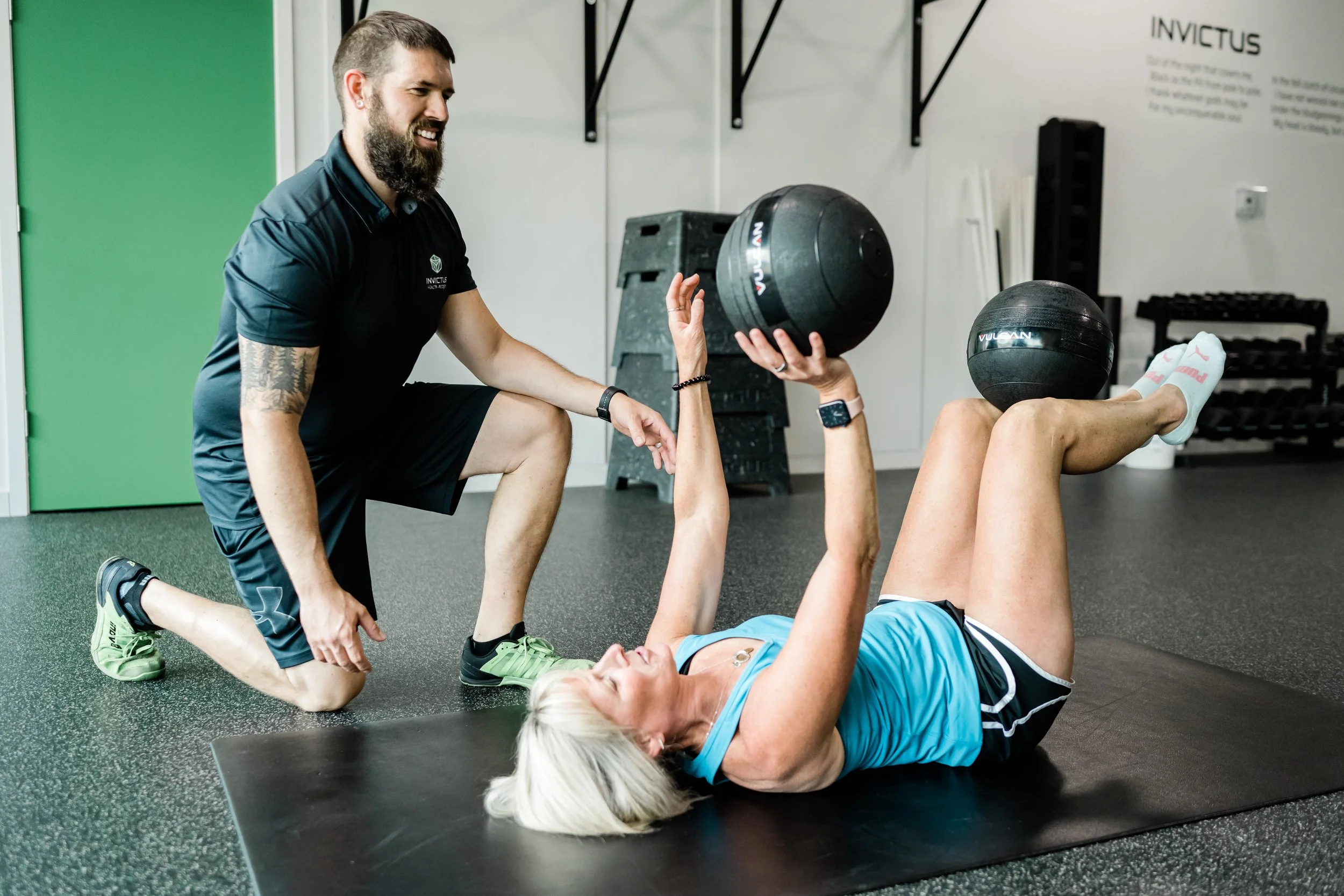 Fitness instructor with a beard assisting a woman doing a leg raise exercise with a medicine ball during a workout session at a gym.