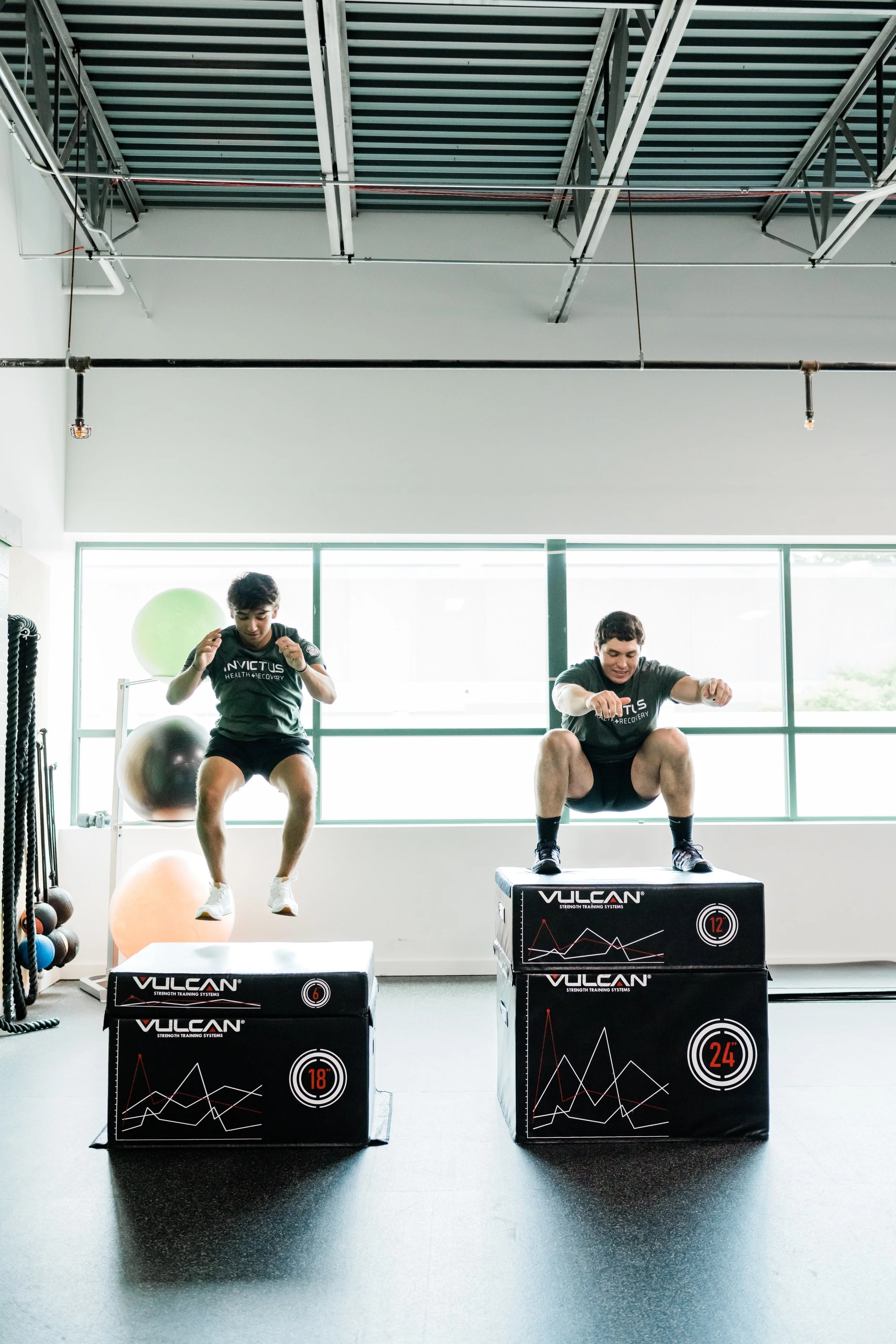 Two men are performing box jumps onto black Vulkan plyometric boxes in a gym. Both men are wearing black t-shirts and shorts, with one on the left mid-air and the other on the right landing on the box. The gym has large windows, exercise balls, and wall-mounted equipment, with a high ceiling and visible piping.