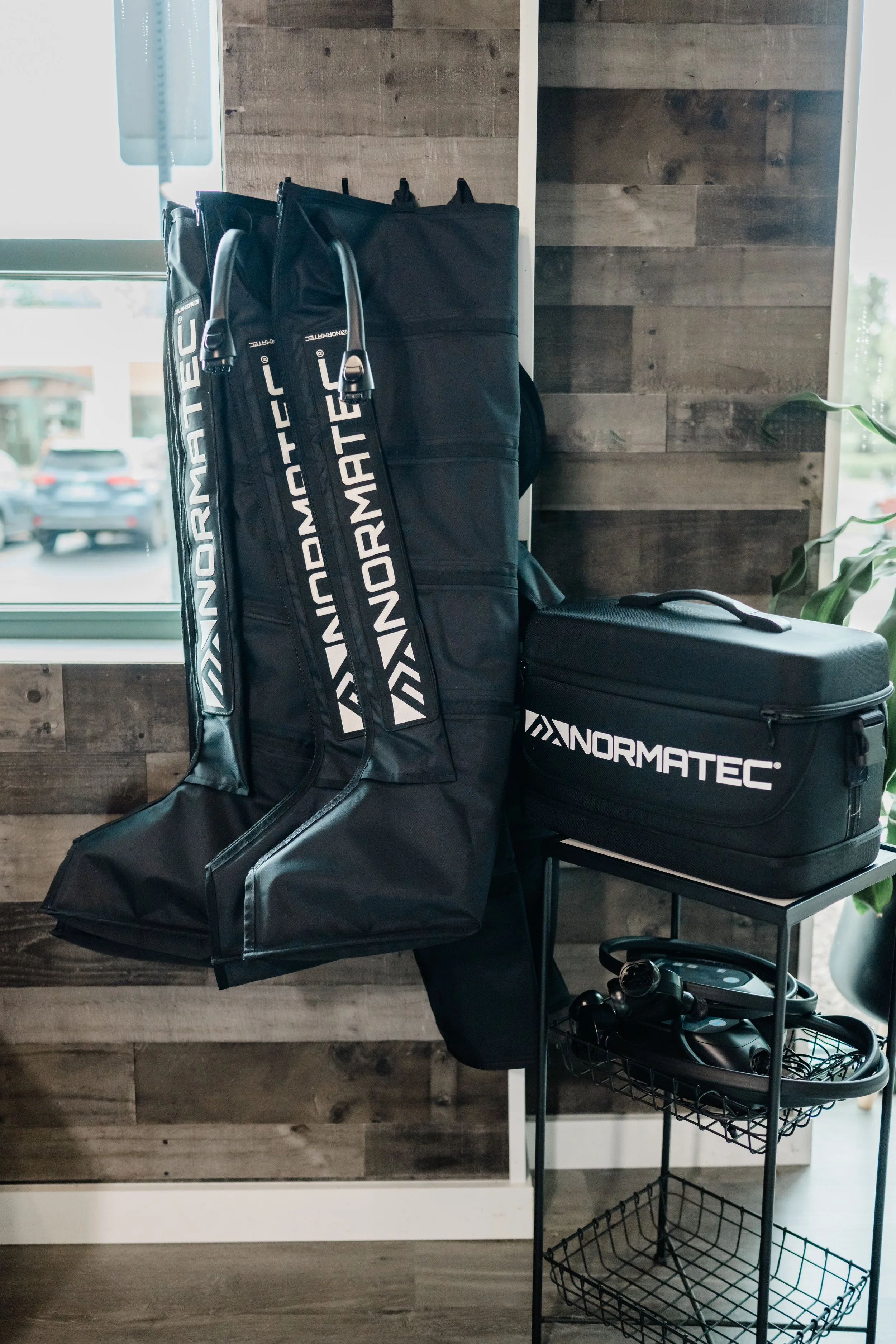 Golf bags with clubs, a black golf club cover, and a black equipment case with 'NORMATEC' printed on it, placed on a black metal storage cart next to a window and a wooden wall.