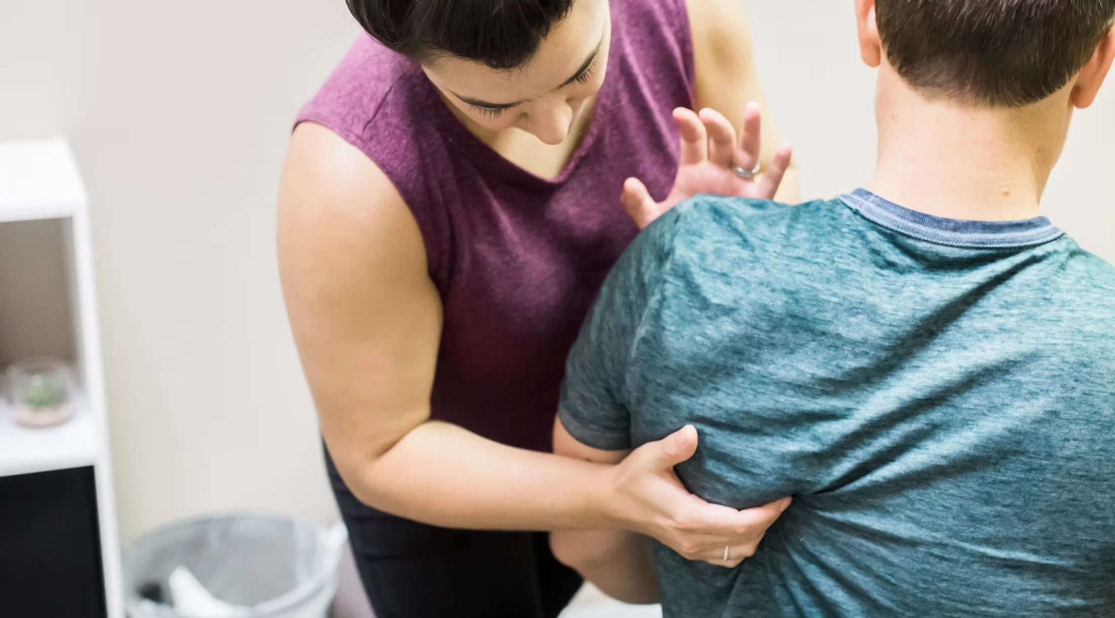 A woman in a purple sleeveless top is being vaccinated by a healthcare worker. The healthcare worker's hands are visible, holding the woman's arm as the injection is administered. The woman's head is bent forward slightly, and she is looking down at her arm. The background is a plain wall, and part of a white shelf with medical supplies can be seen on the left side of the image.