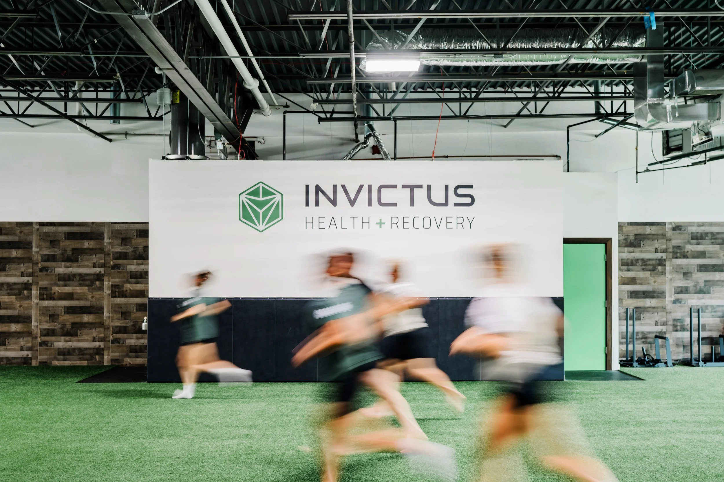 Blurred people running indoors in front of a wall with a sign that reads 'Invictus Health + Recovery.'