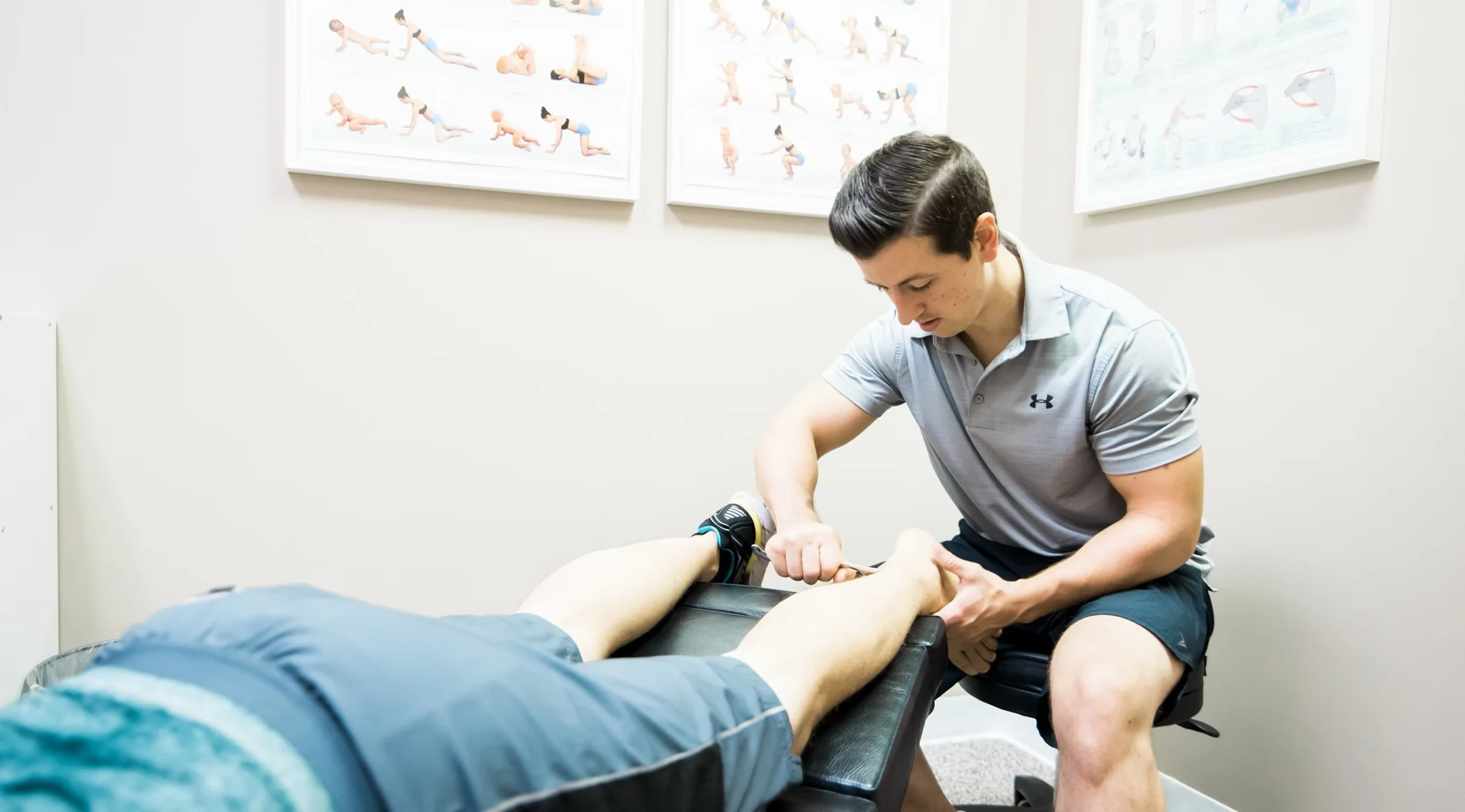 A physical therapist is kneeling beside a patient lying face down on a treatment table, performing a lower-leg manual therapy or assessment. The room has posters on exercise and anatomy on the wall.