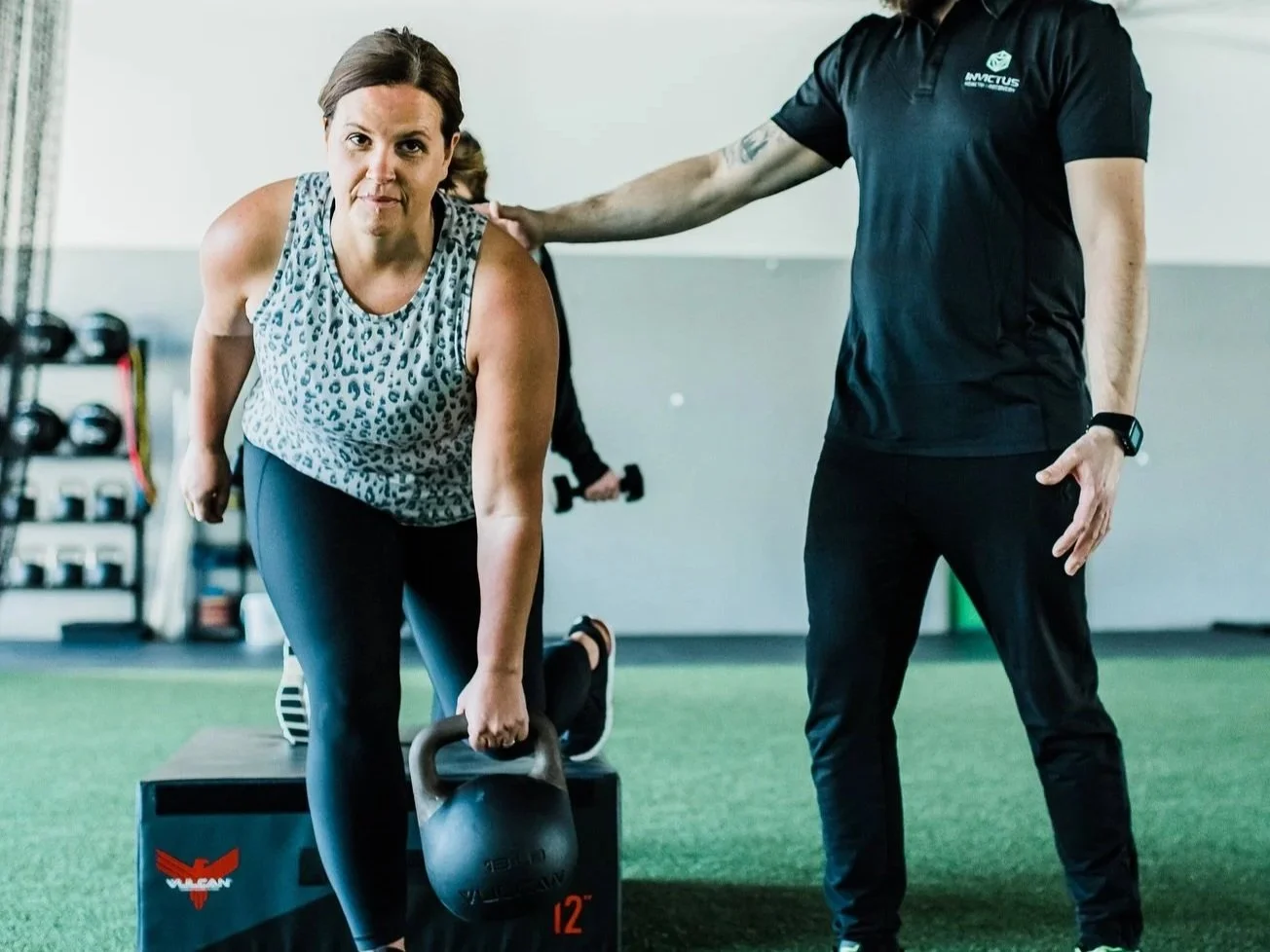 woman performing a kettlebell exercise with personal trainer coaching her in a gym.