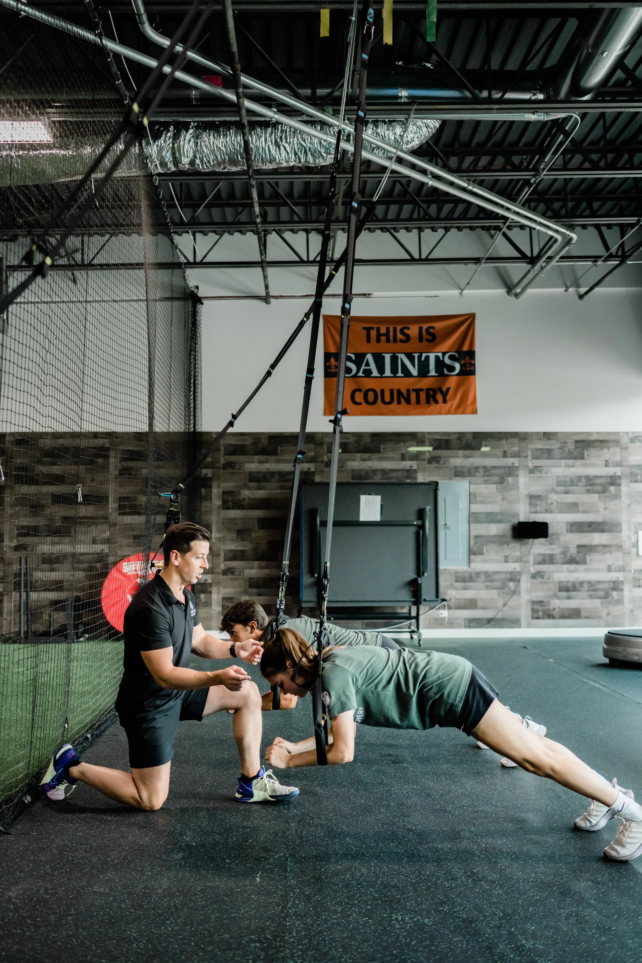 A fitness trainer assisting a woman during a plank exercise in a gym with a sign that reads 'This is Saints Country' on the wall.