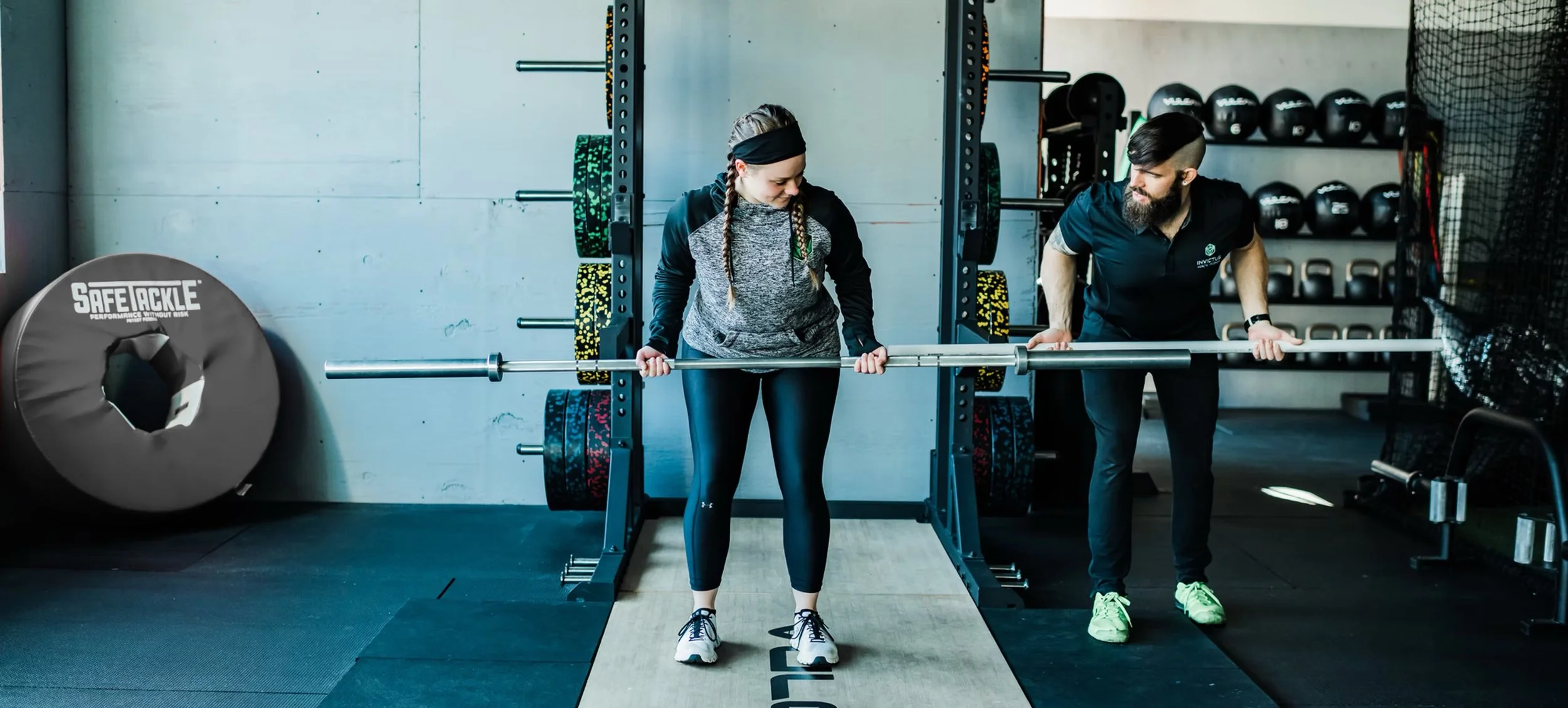 A fitness trainer assists a woman with a barbell during weight training in a gym. The woman is preparing to lift, and both are focused on the barbell.