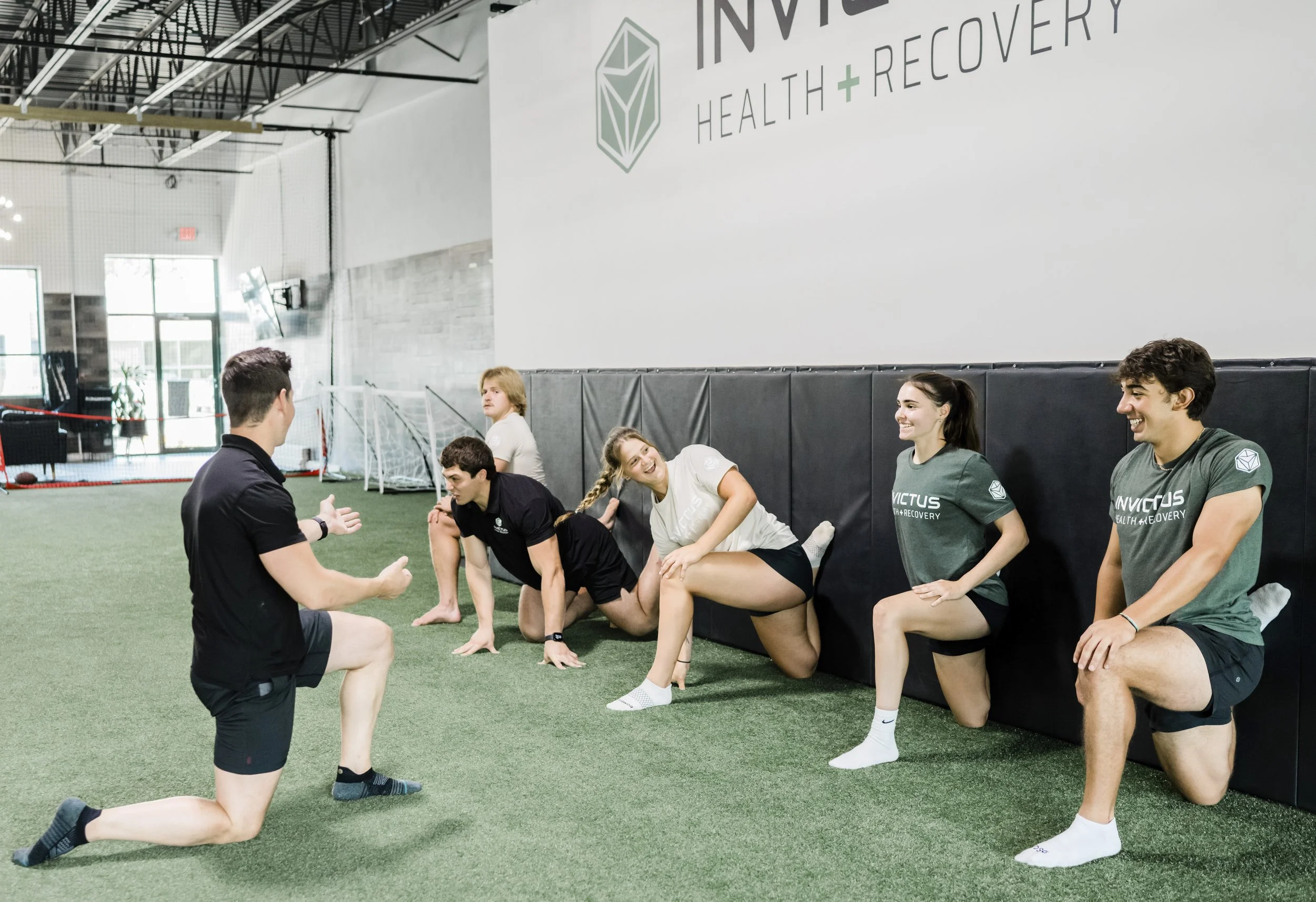 Group of people sitting on the floor in sportswear, engaging in a conversation during a fitness or rehabilitation session inside a gym or sports facility.
