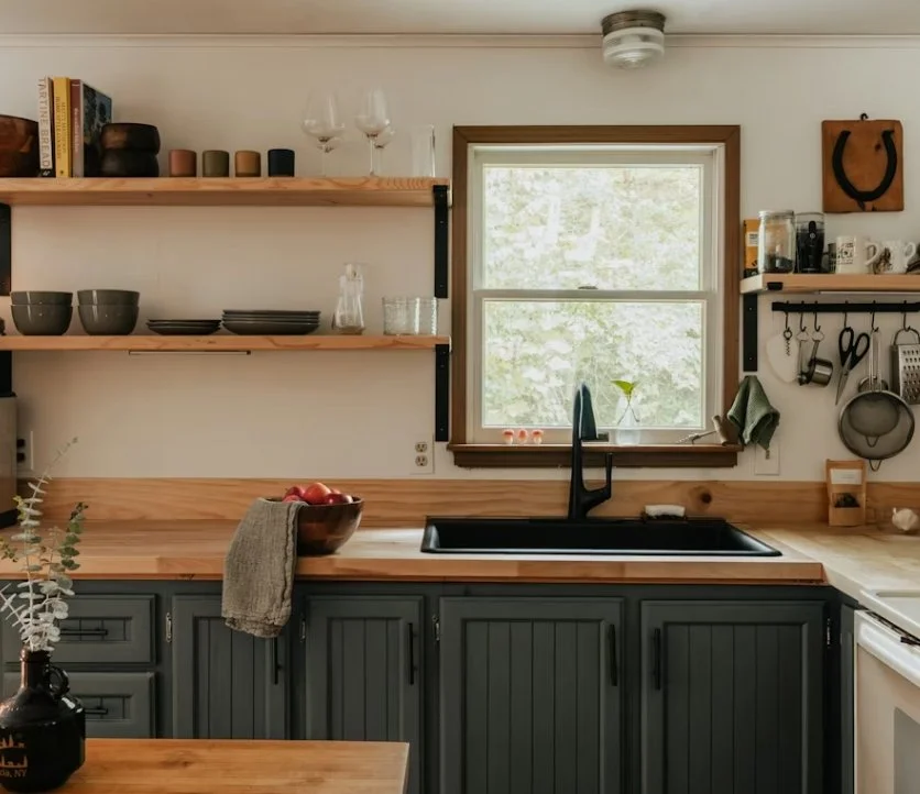 Kitchen with a wooden countertop, dark green cabinets, open shelves with dishes, a window with greenery outside, and various kitchen utensils and decorations.