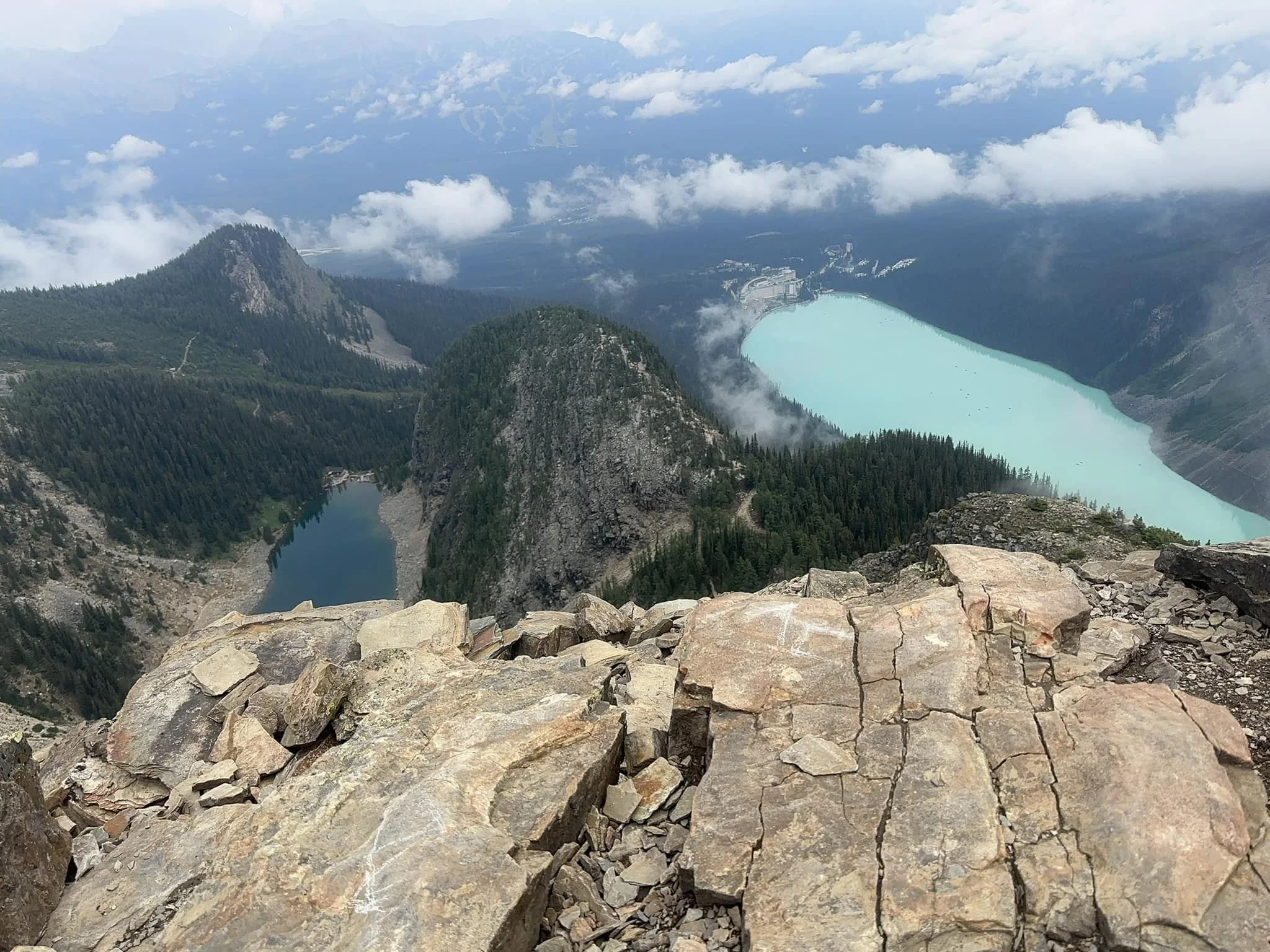 View from a mountain top overlooking a lake with turquoise water, surrounded by forested slopes and rocky outcrops, with clouds overhead.
