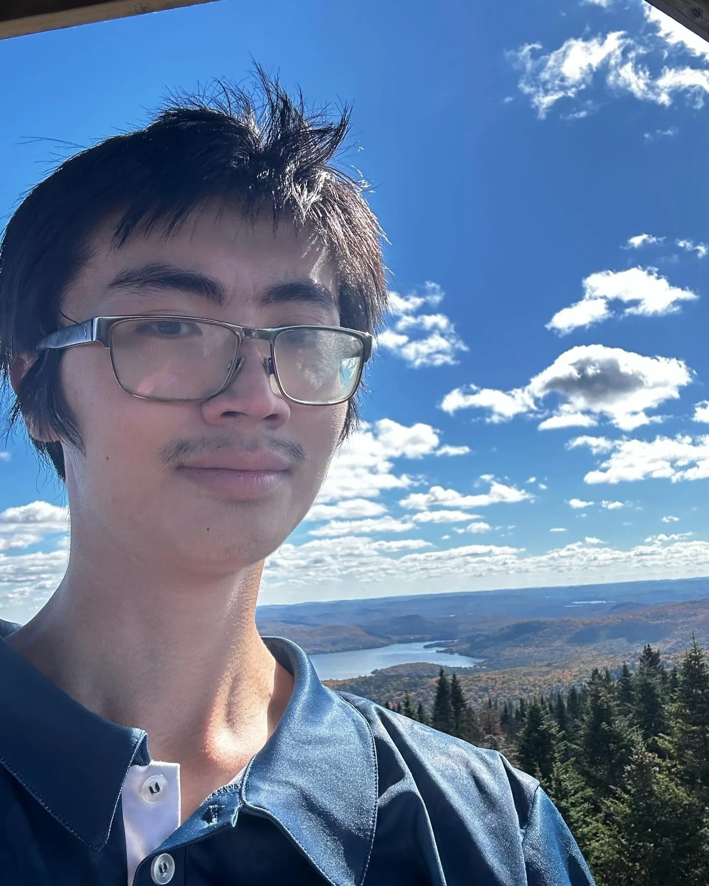 A young man with glasses taking a selfie outdoors with a scenic view of a river, hills, and a partly cloudy blue sky in the background.