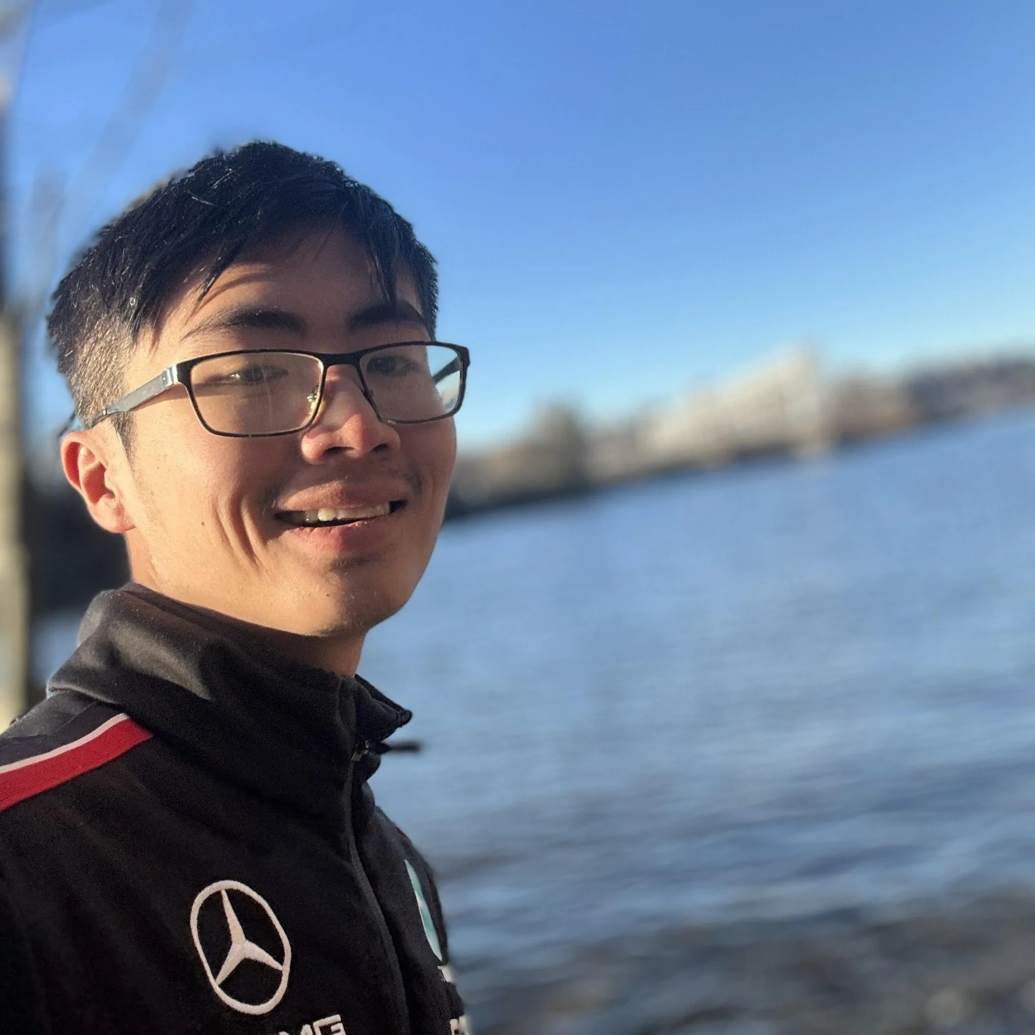 Close-up of a person's face with glasses, smiling, outdoors near water on a sunny day.