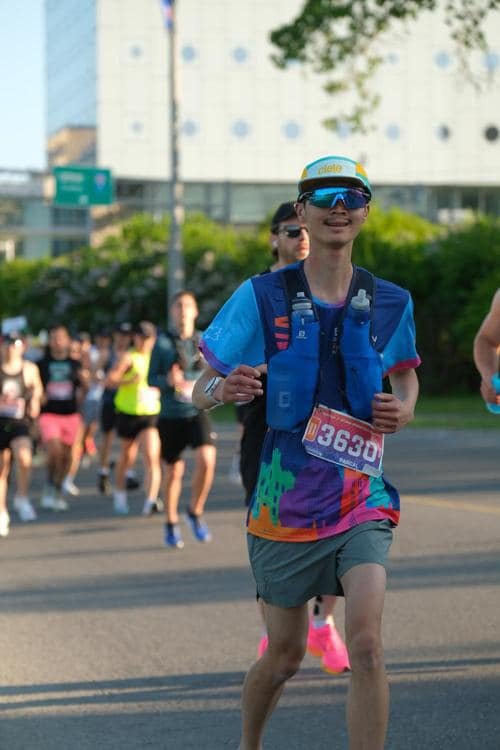 A young man running in a marathon, wearing sunglasses, a blue hydration vest, a colorful shirt, gray shorts, and a race bib number 3620.