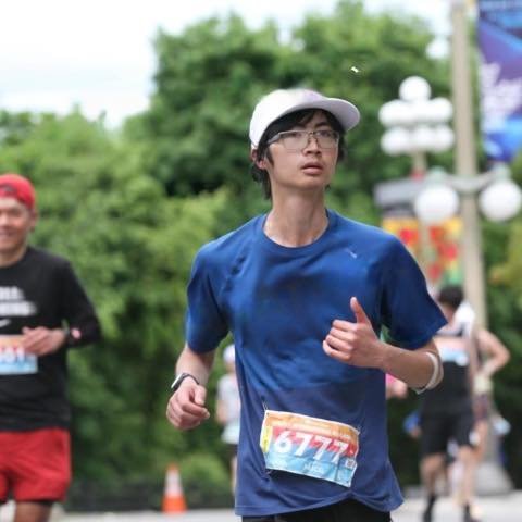 A young man wearing glasses, a white cap, and a blue t-shirt running in a race with a bib number, in a park or outdoor setting with other runners and trees in the background.