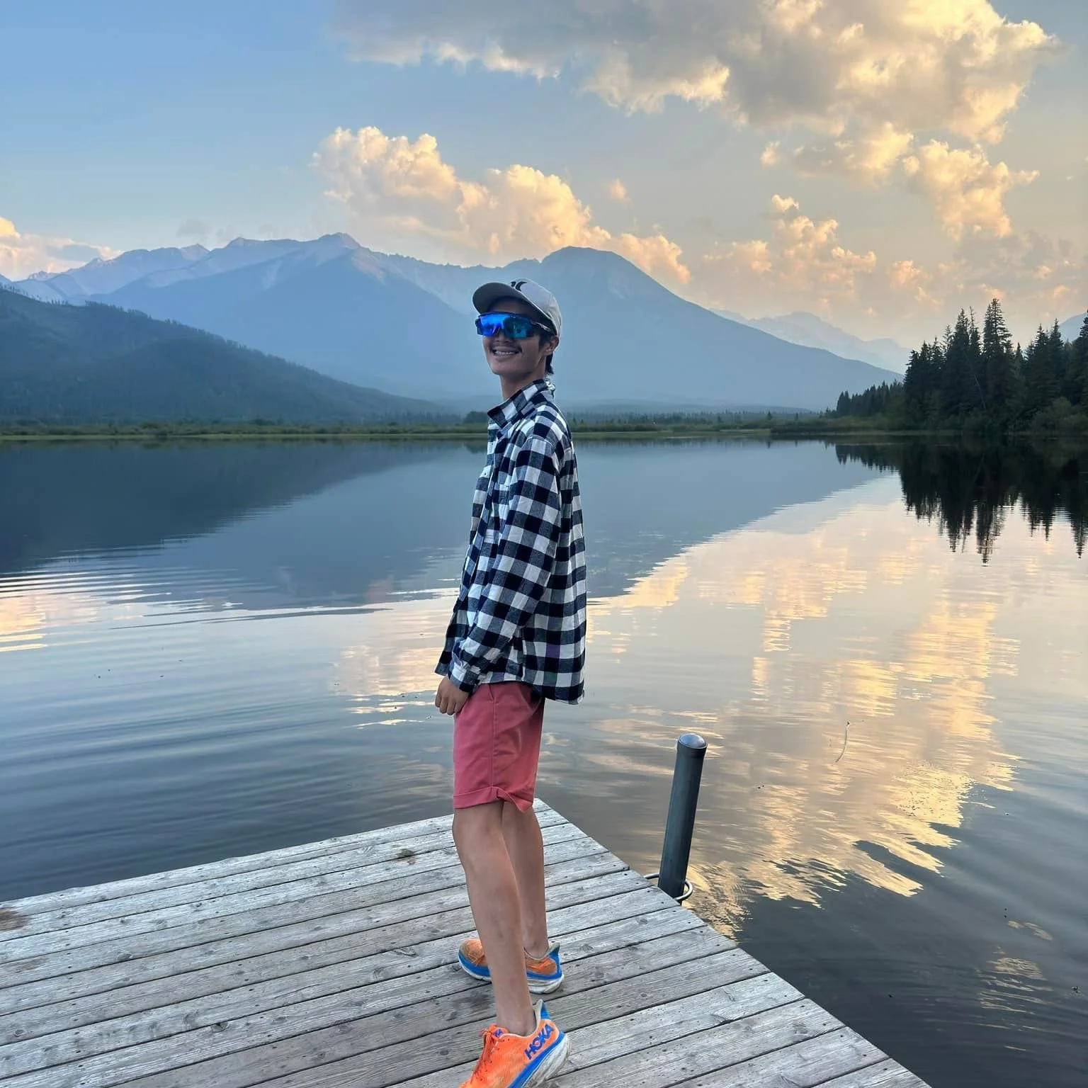 Person standing on a wooden dock beside a calm lake, with mountains and a partly cloudy sky in the background.