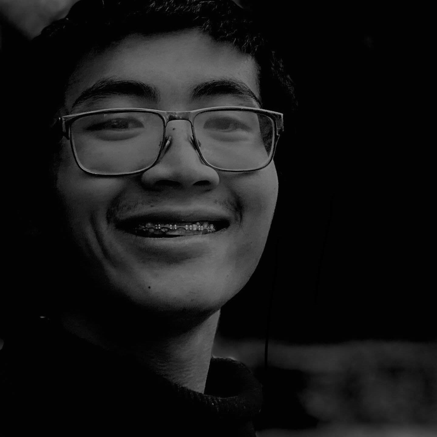 A smiling young person wearing glasses and braces, in black and white, with a dark background.