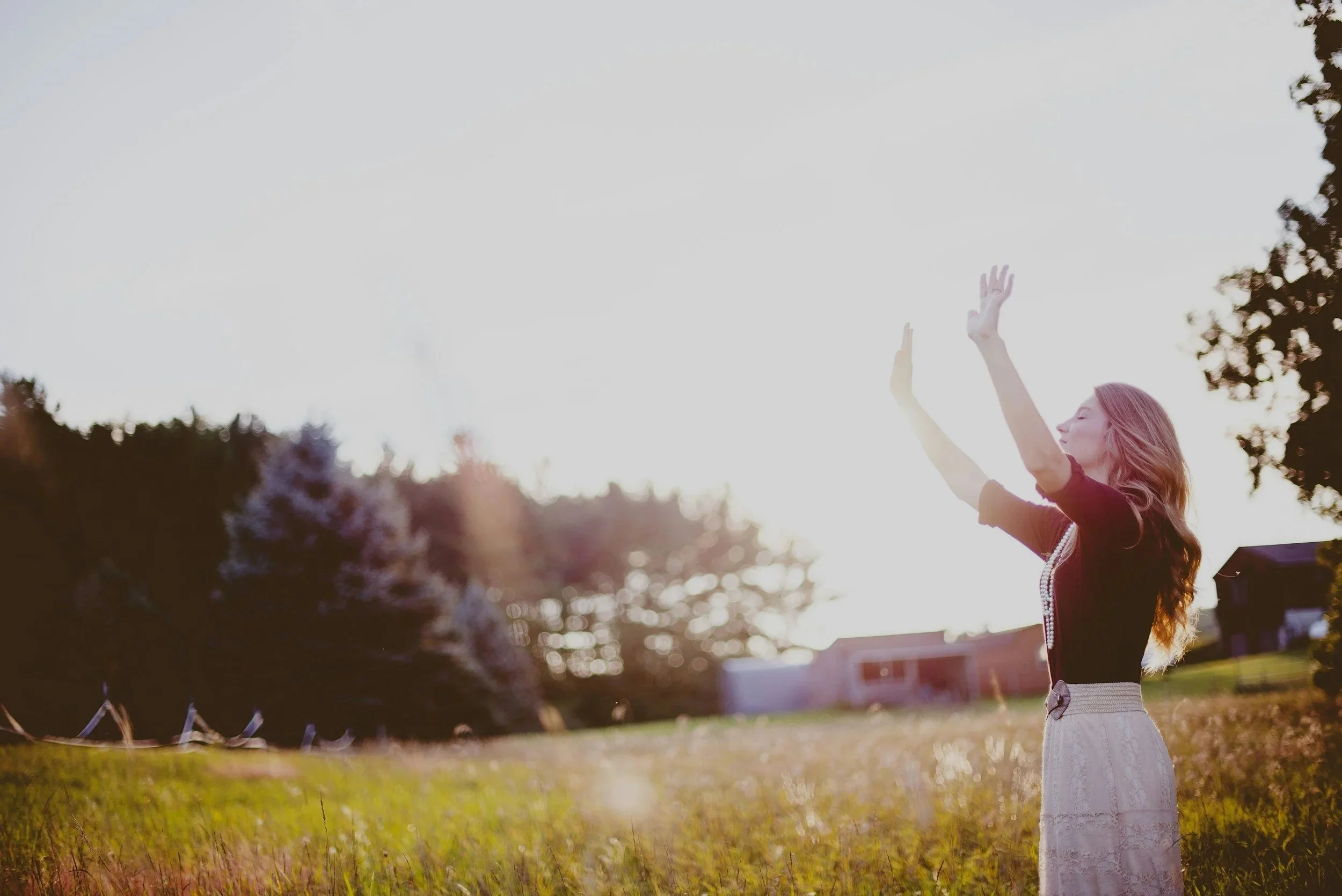 A woman with long hair wearing a black top and white skirt stands outdoors in a grassy area, raising her hands towards the sky as the sun sets or rises, with a background of trees and a clear sky.