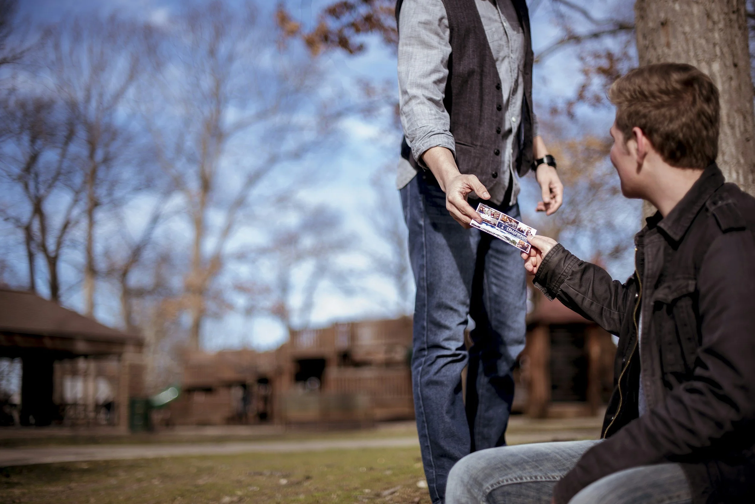 A person standing outdoors in a park handing a photo strip to a person sitting on the ground. There are trees with mostly bare branches and a blue sky in the background.