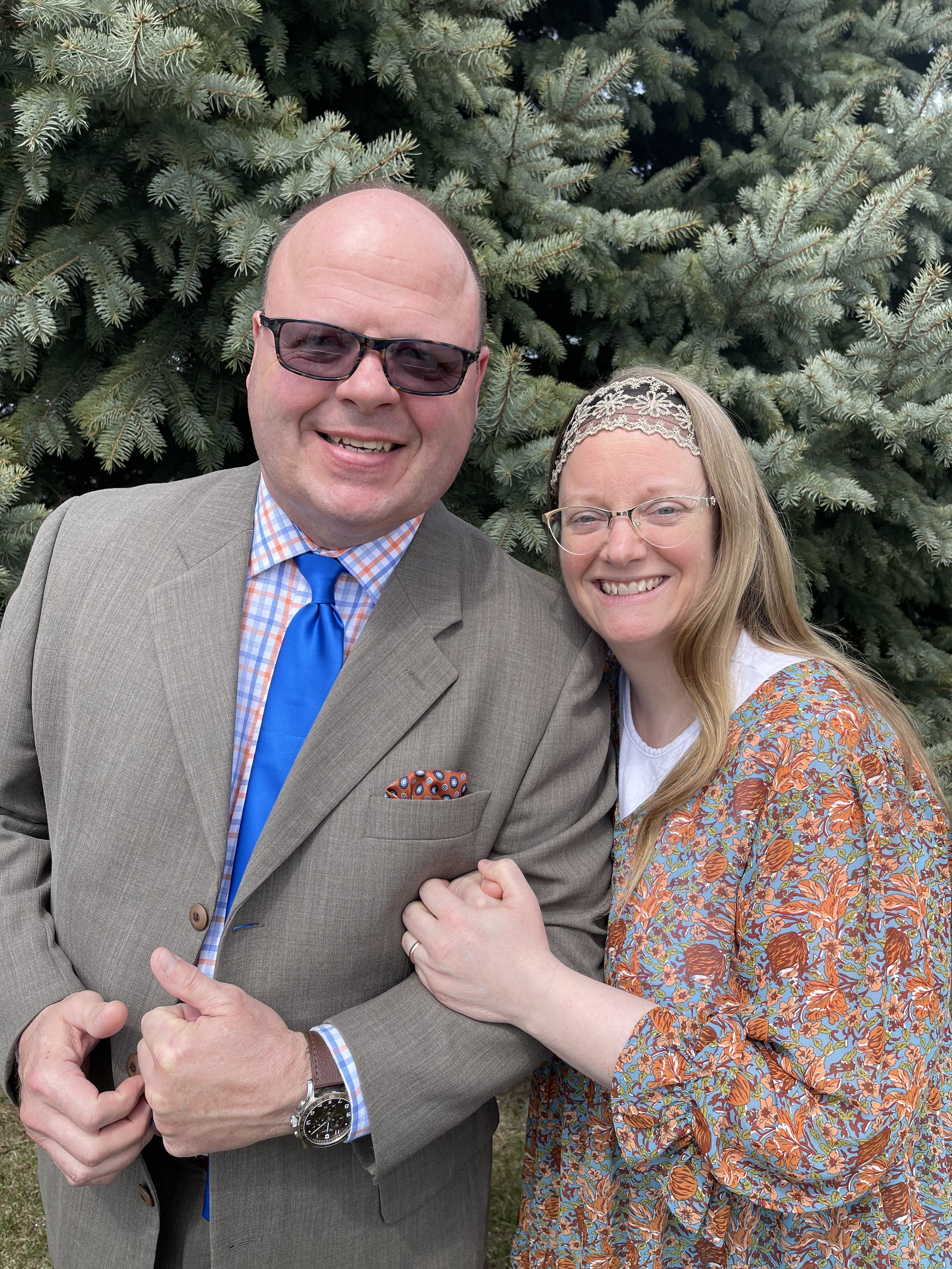 A man in a gray suit, blue tie, and sunglasses, and a woman with long hair, glasses, and a floral patterned dress, smiling and holding hands in front of a pine tree.
