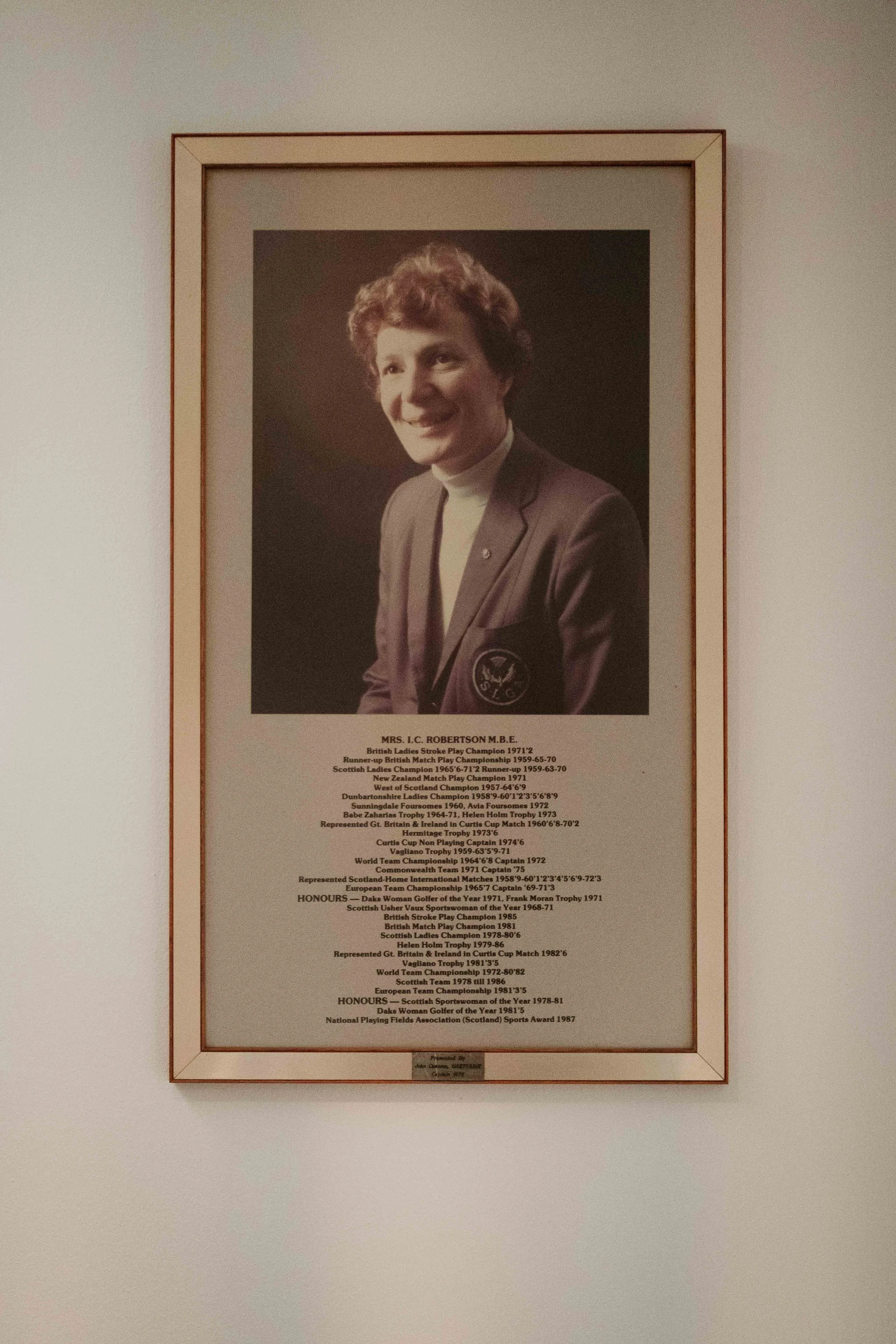 A framed collage of a woman’s portrait and a list of her achievements and honors, displayed on a white wall.