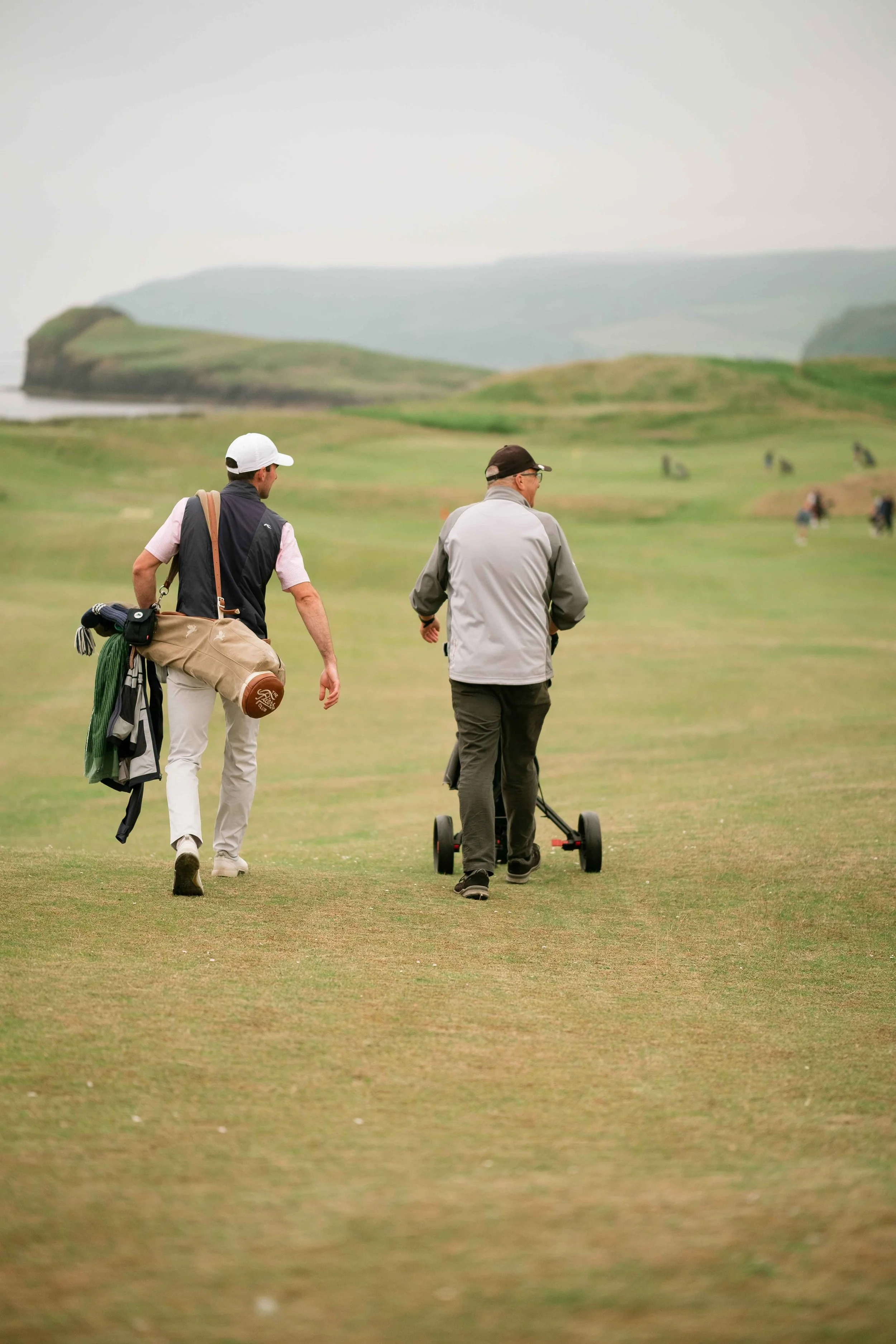 Two men walking on a golf course, one pushing a golf trolley, with rolling green hills and scattered trees in the background.
