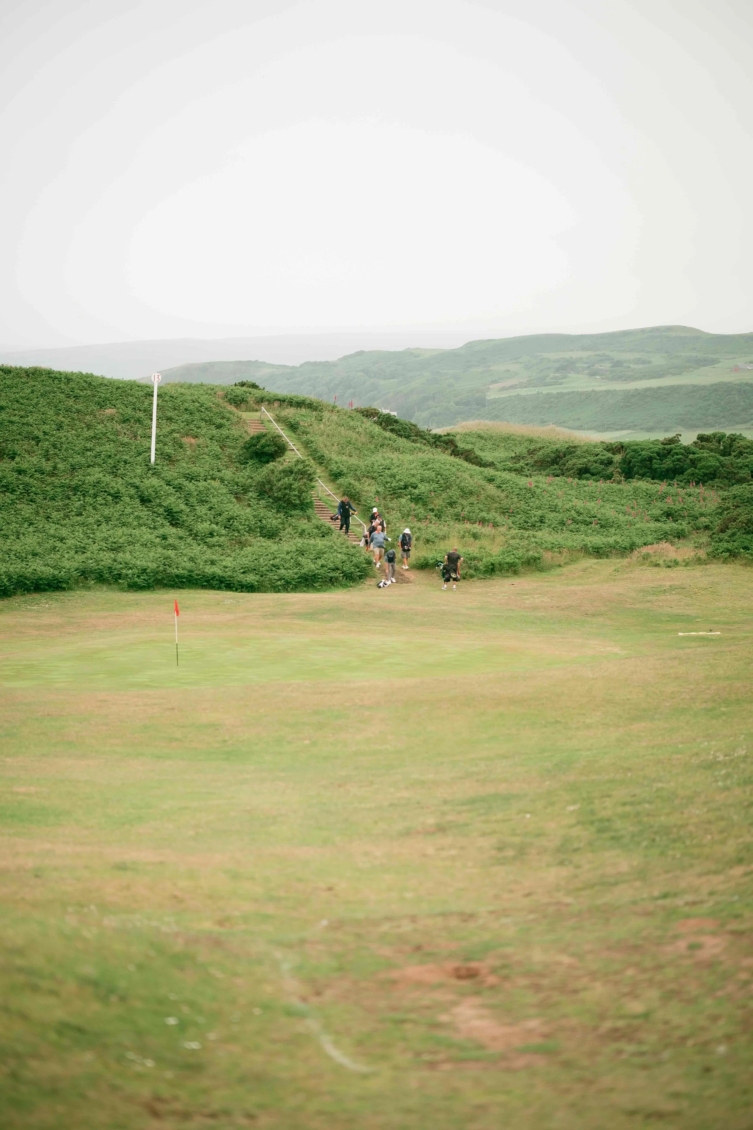 Golf course surrounded by green hills with a group of people walking up stairs and a few standing, overcast sky.