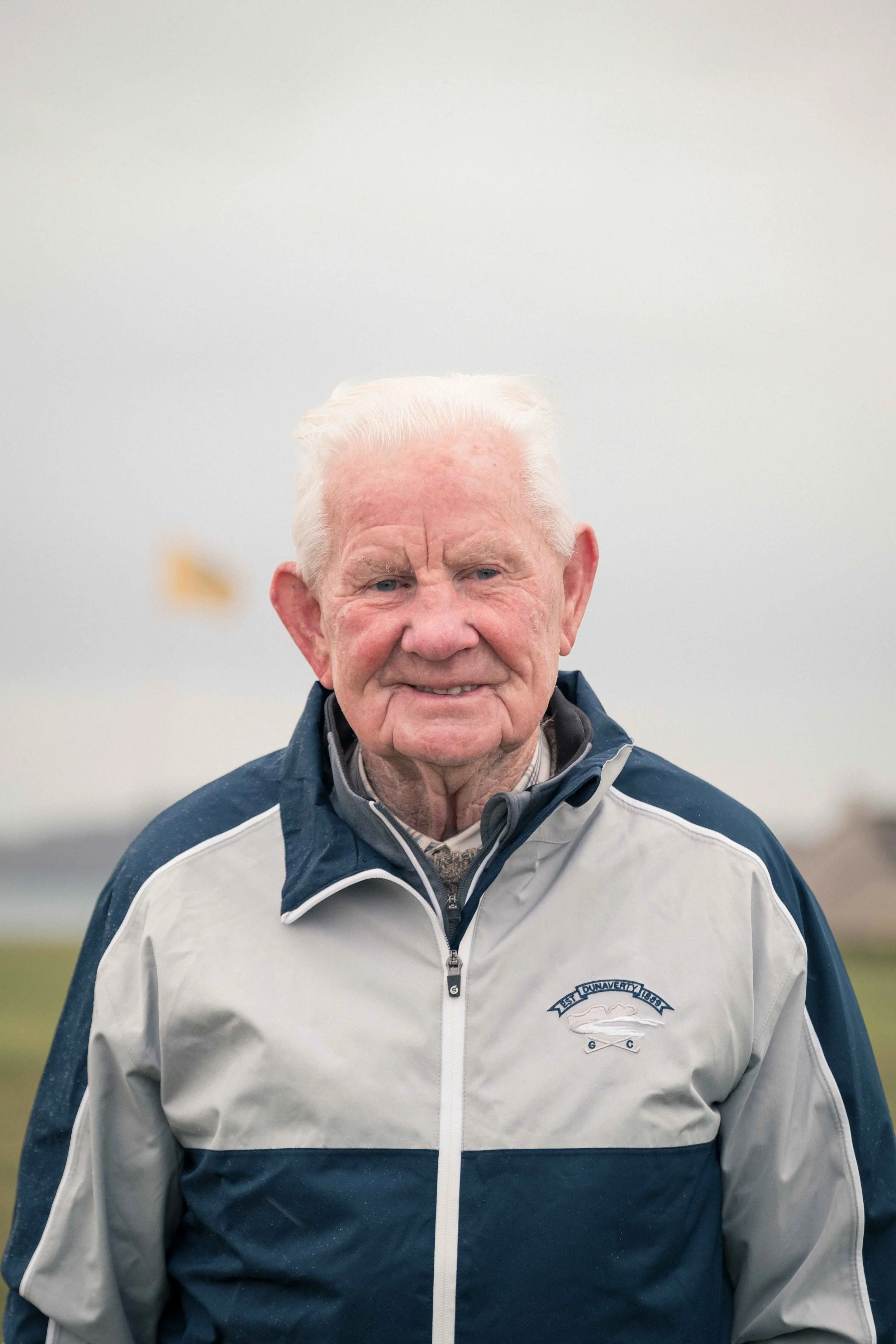An elderly man with white hair and a friendly smile, wearing a navy blue and white jacket with a logo, standing outdoors against a cloudy sky.