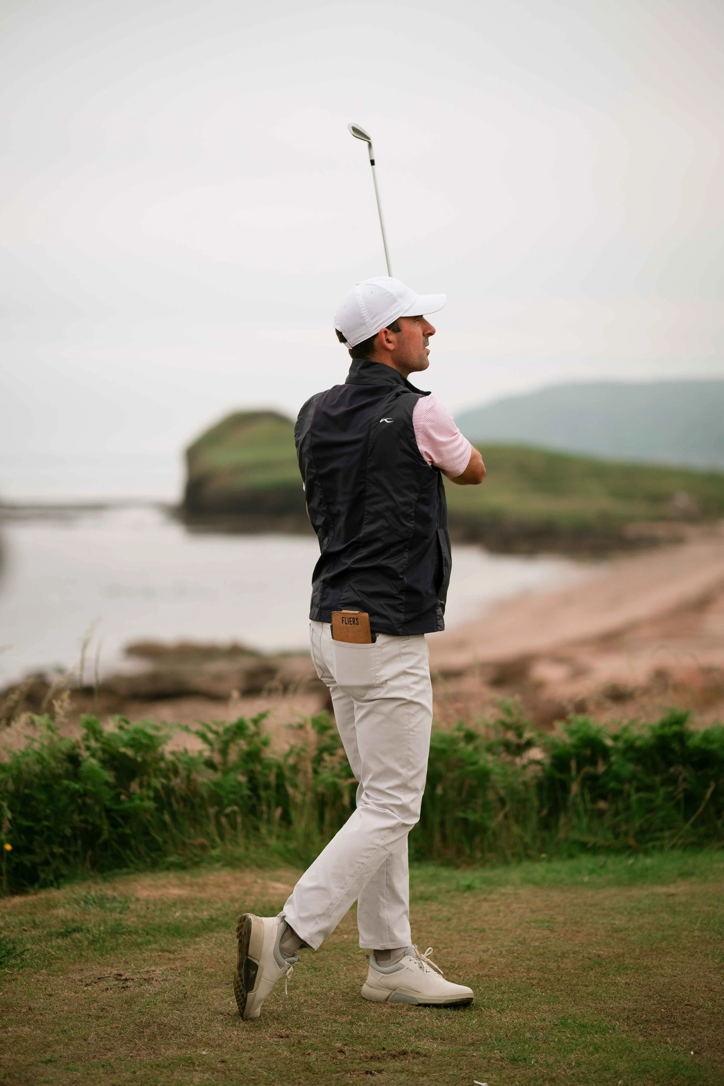 A man playing golf on a golf course, standing on the grass with a golf club balanced on his head, dressed in a white cap, black vest, pink shirt, and light-colored pants.
