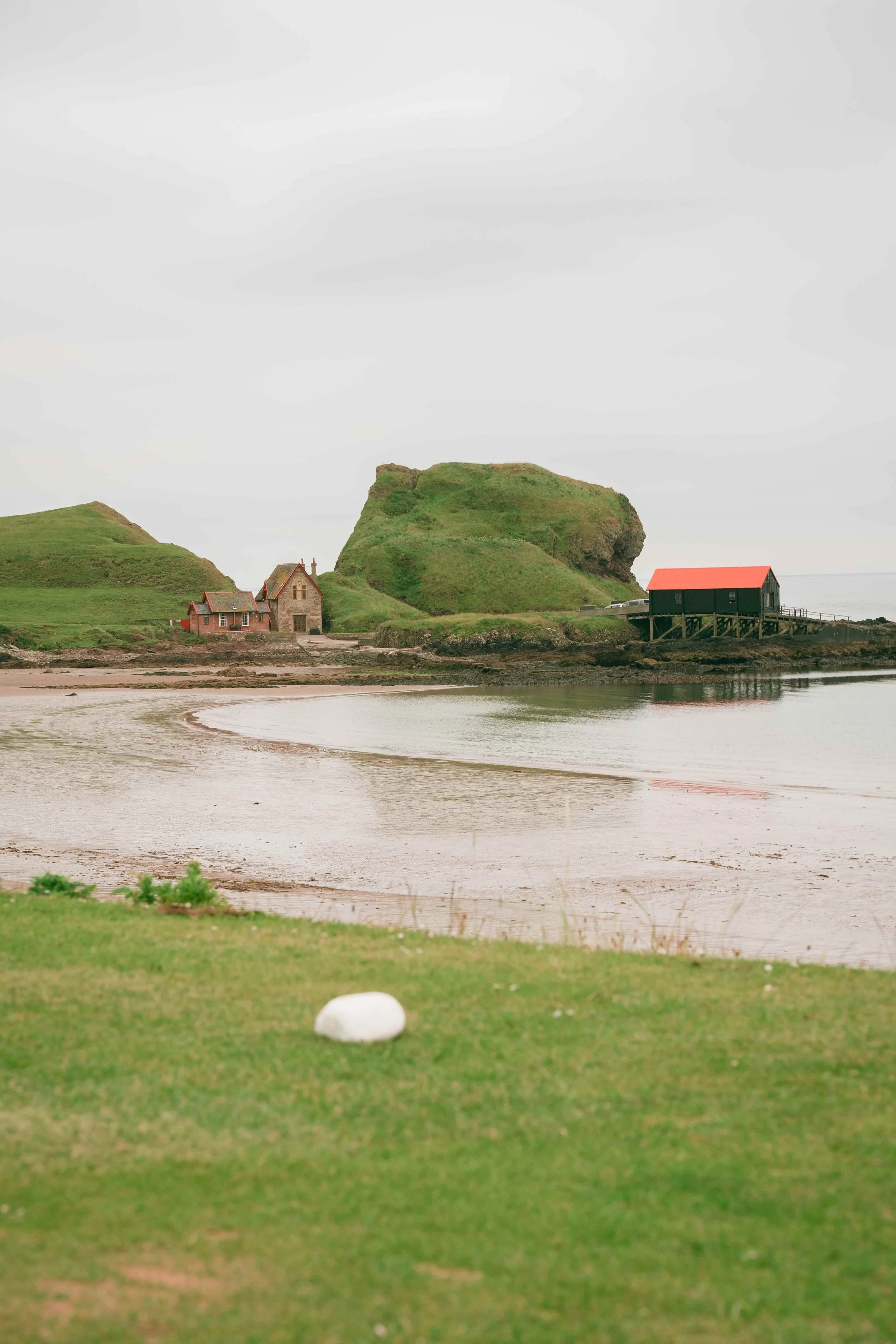 A coastal scene with grassy hills, a beach, and pavilions. A large, rocky hill in the background with a person walking along the shore.