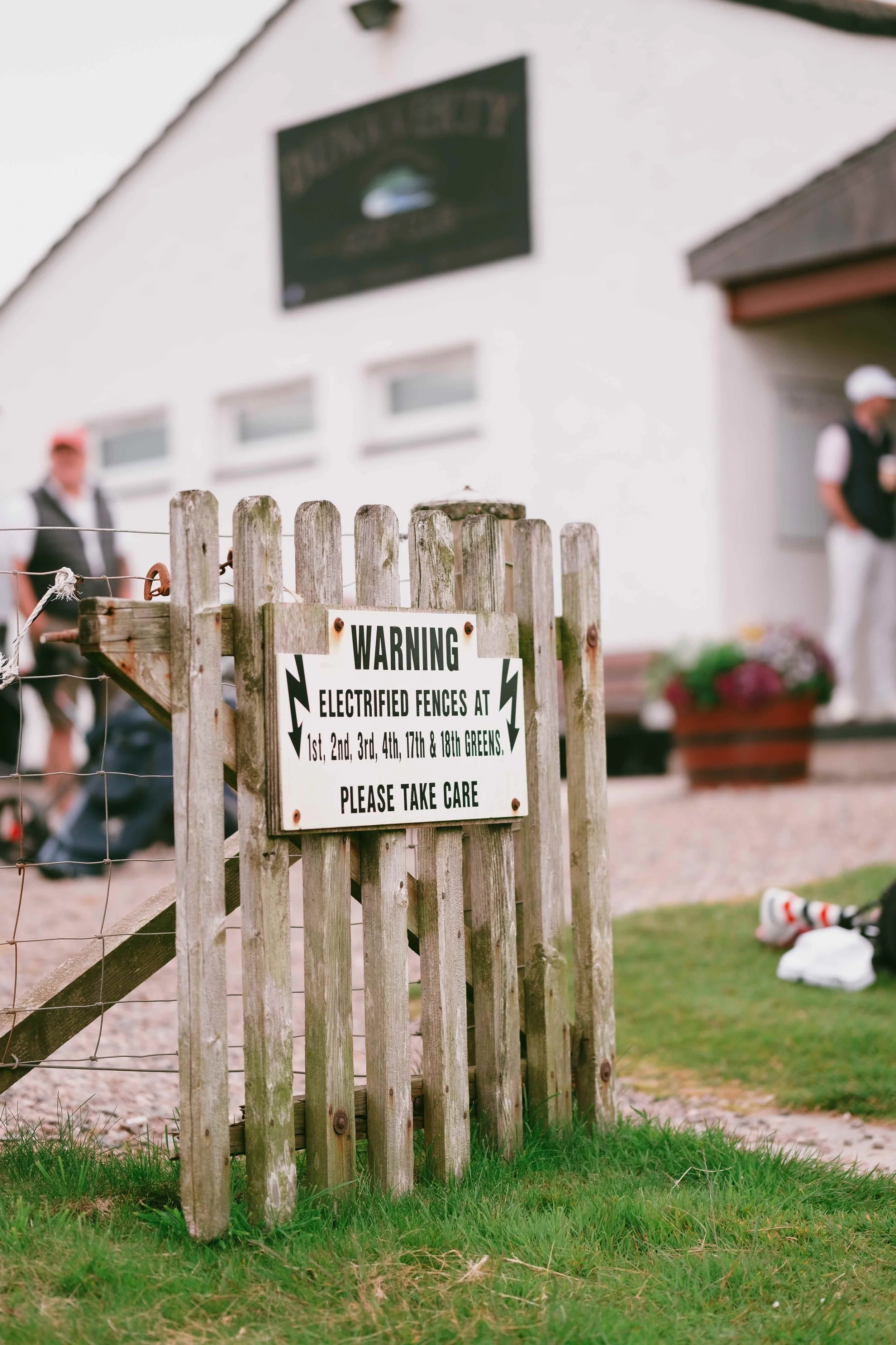 A wooden gate with a warning sign about electric fences, located outside a golf club building. In the background, people are standing near the building, and a potted plant is visible. The building has a sign that reads "Golf Club".